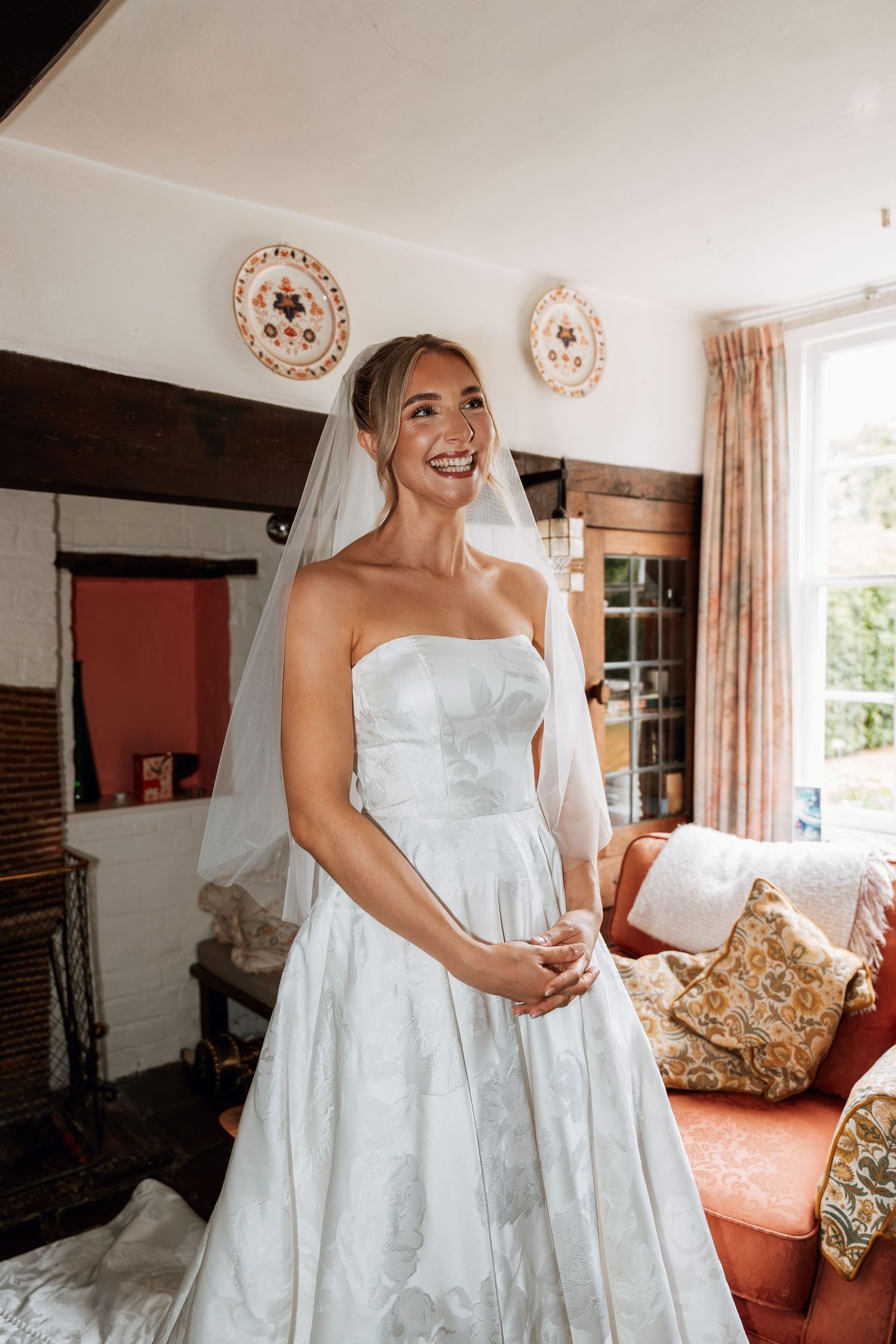 A strapless white wedding gown is showcased in a bright interior room with floral decor and cream curtains.