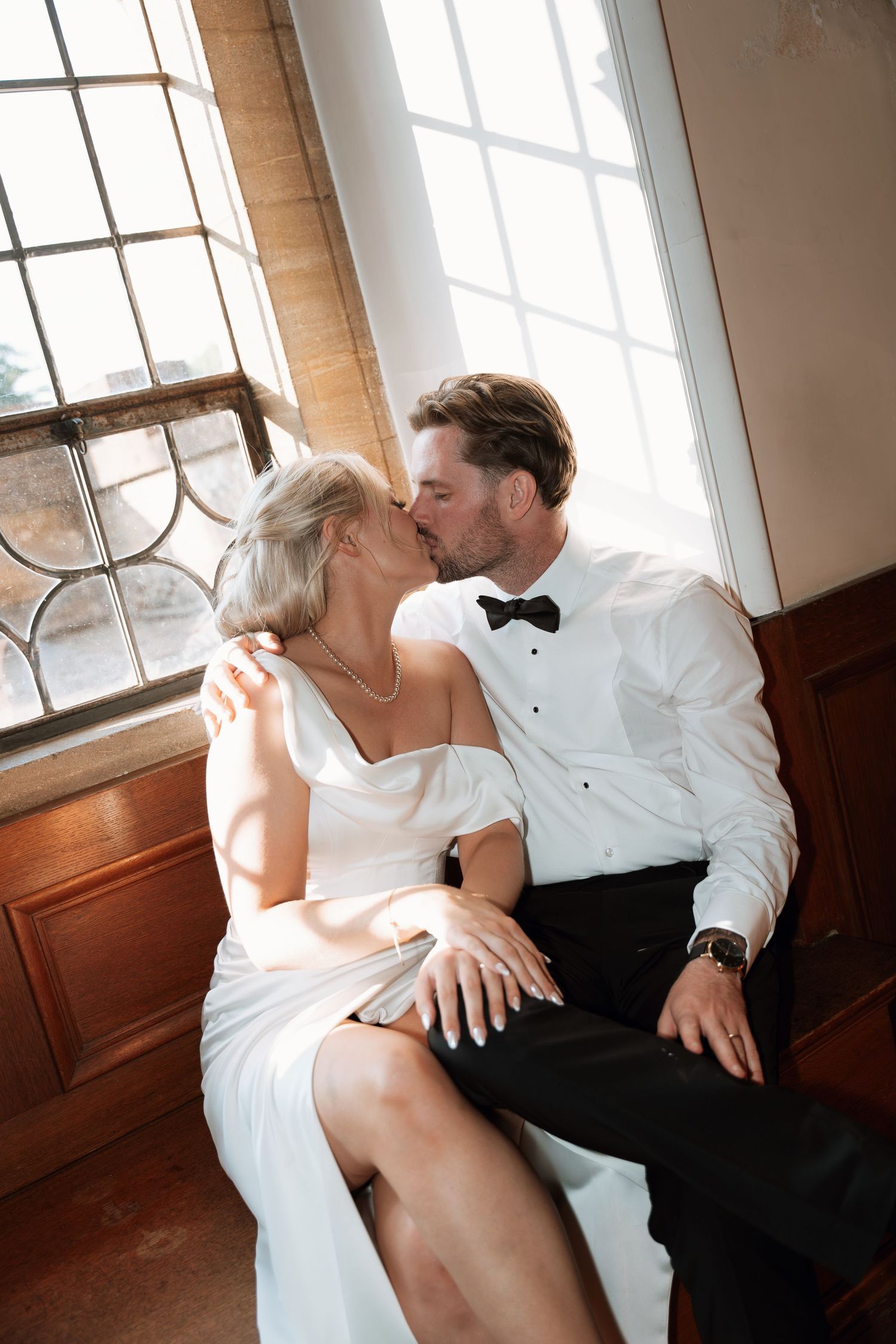 Romantic wedding couple sharing intimate moments on a bench by a window in classic black and white tones.