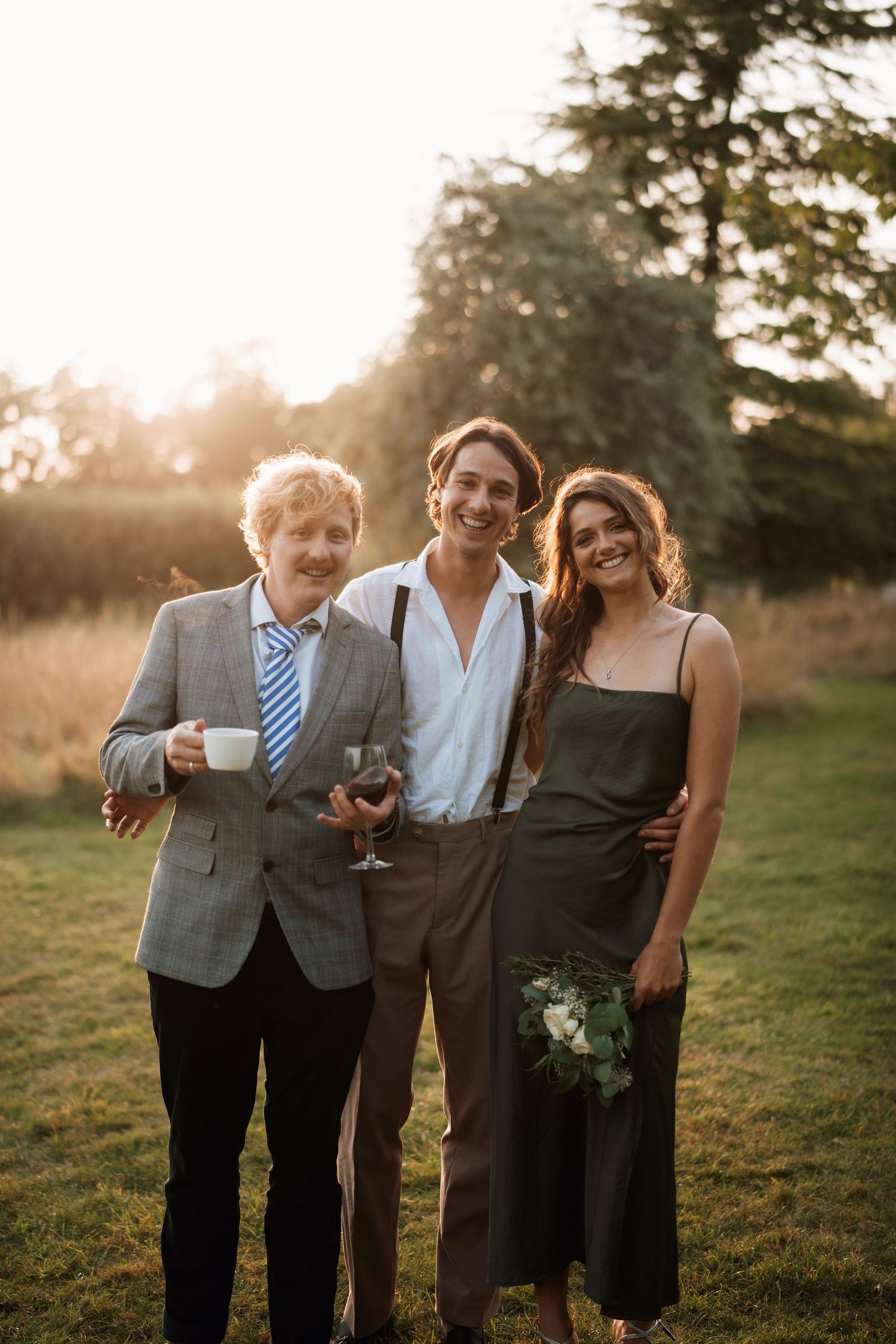 Three wedding guests enjoy drinks and conversation during golden hour at an outdoor reception.