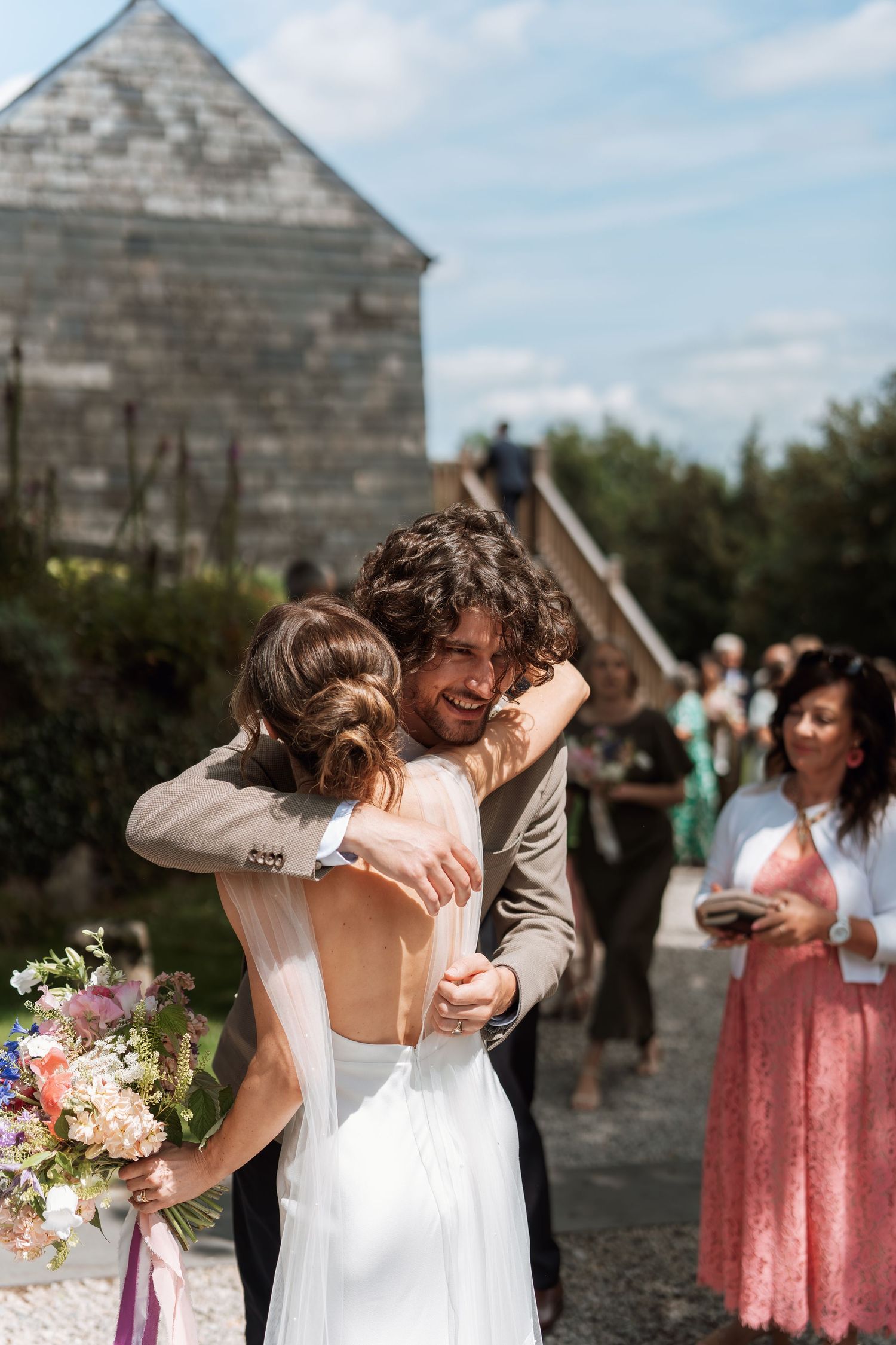 A tender embrace during an outdoor wedding ceremony with guests looking on and a rustic building in the background.