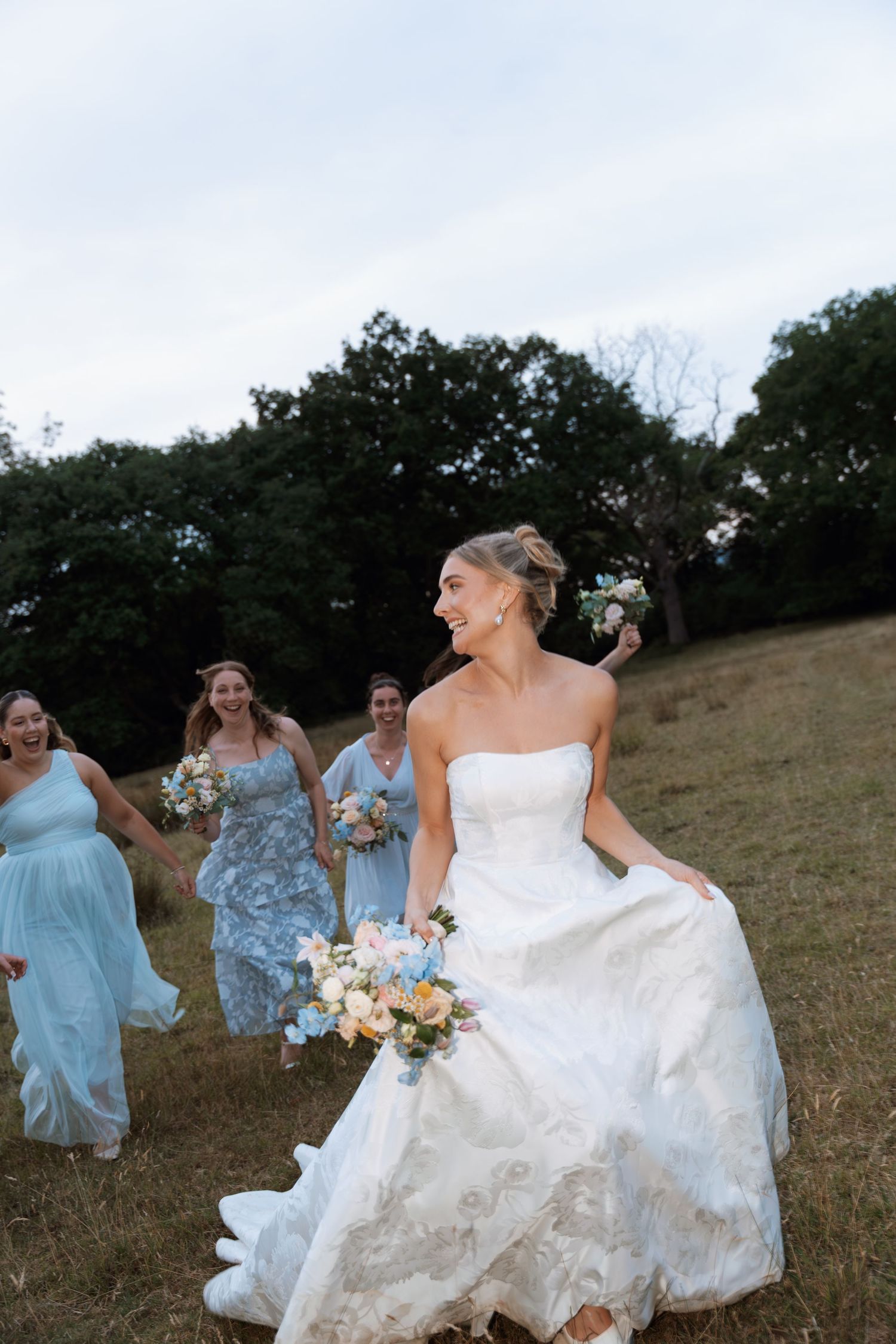 A bride in a flowing white gown walks across a grassy hillside with bridesmaids in blue dresses behind her.