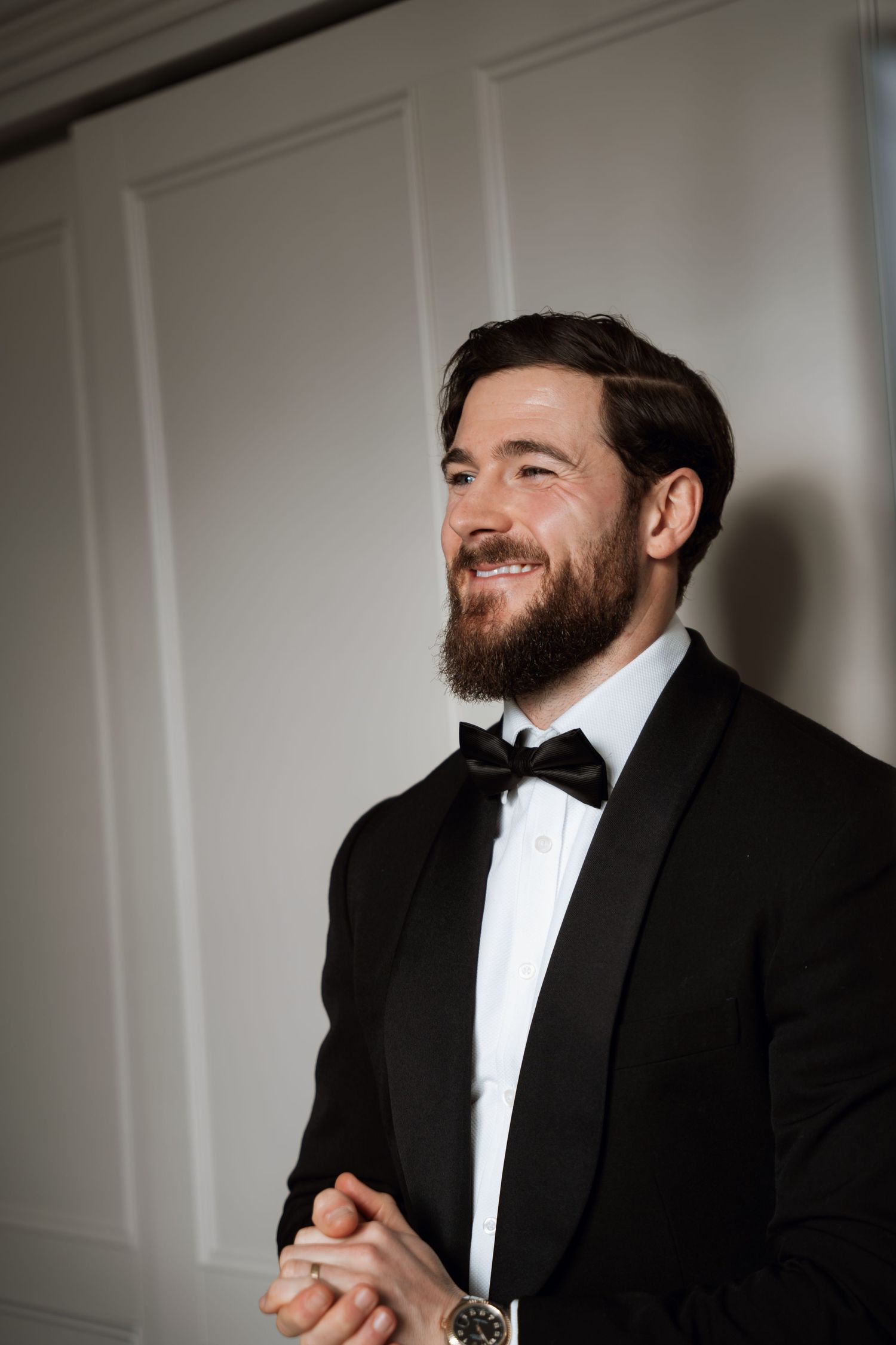 Groom in classic black tuxedo smiling warmly against white wall.