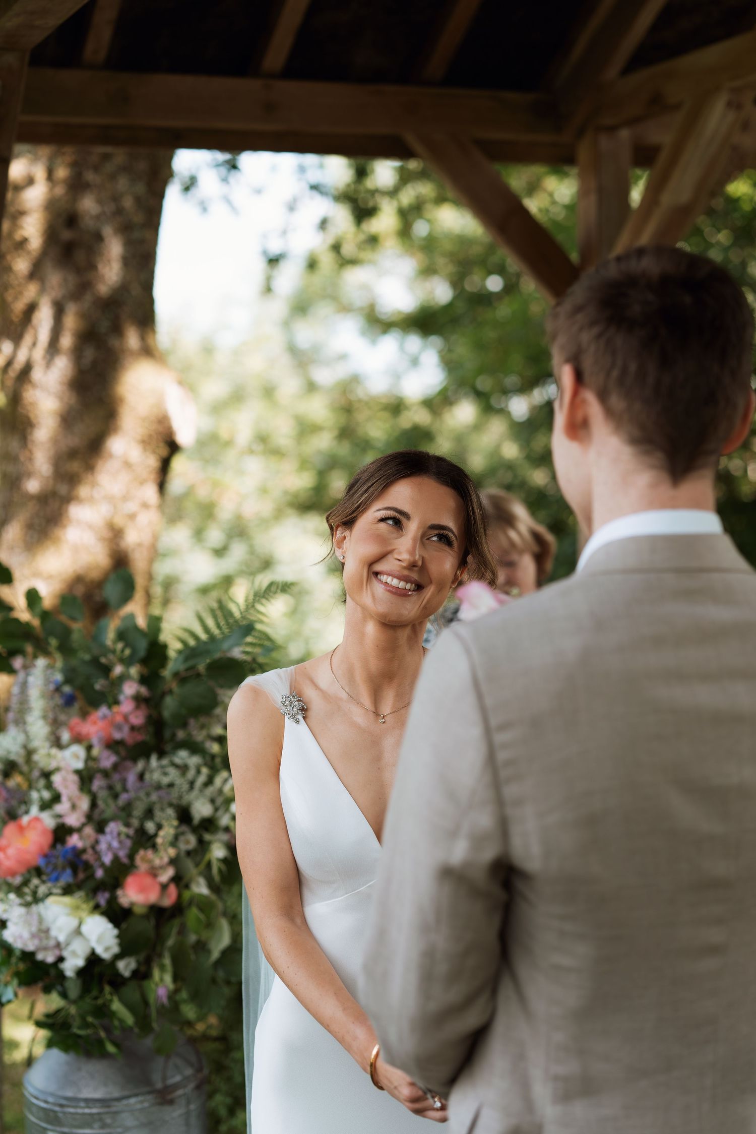 A bride in a sleek white dress smiles during an outdoor wedding ceremony surrounded by flowers.