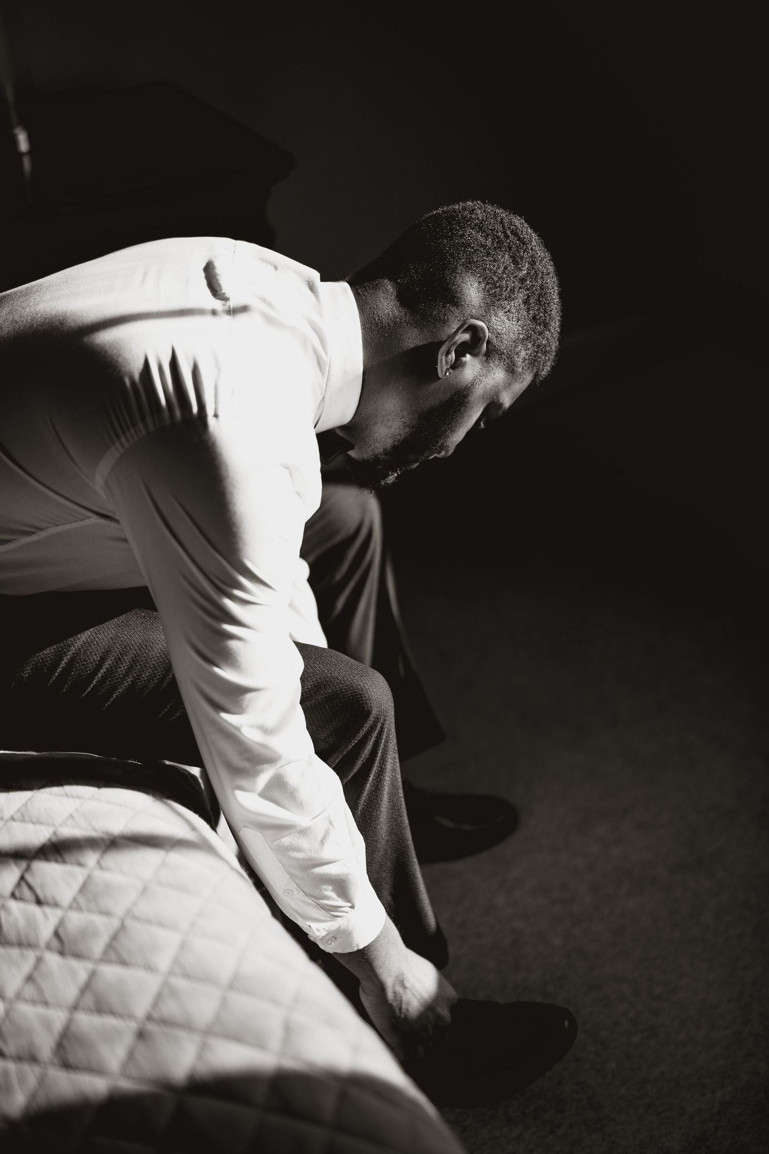 Black and white portrait of a person in a white shirt bending down near a quilted mattress or cushion.