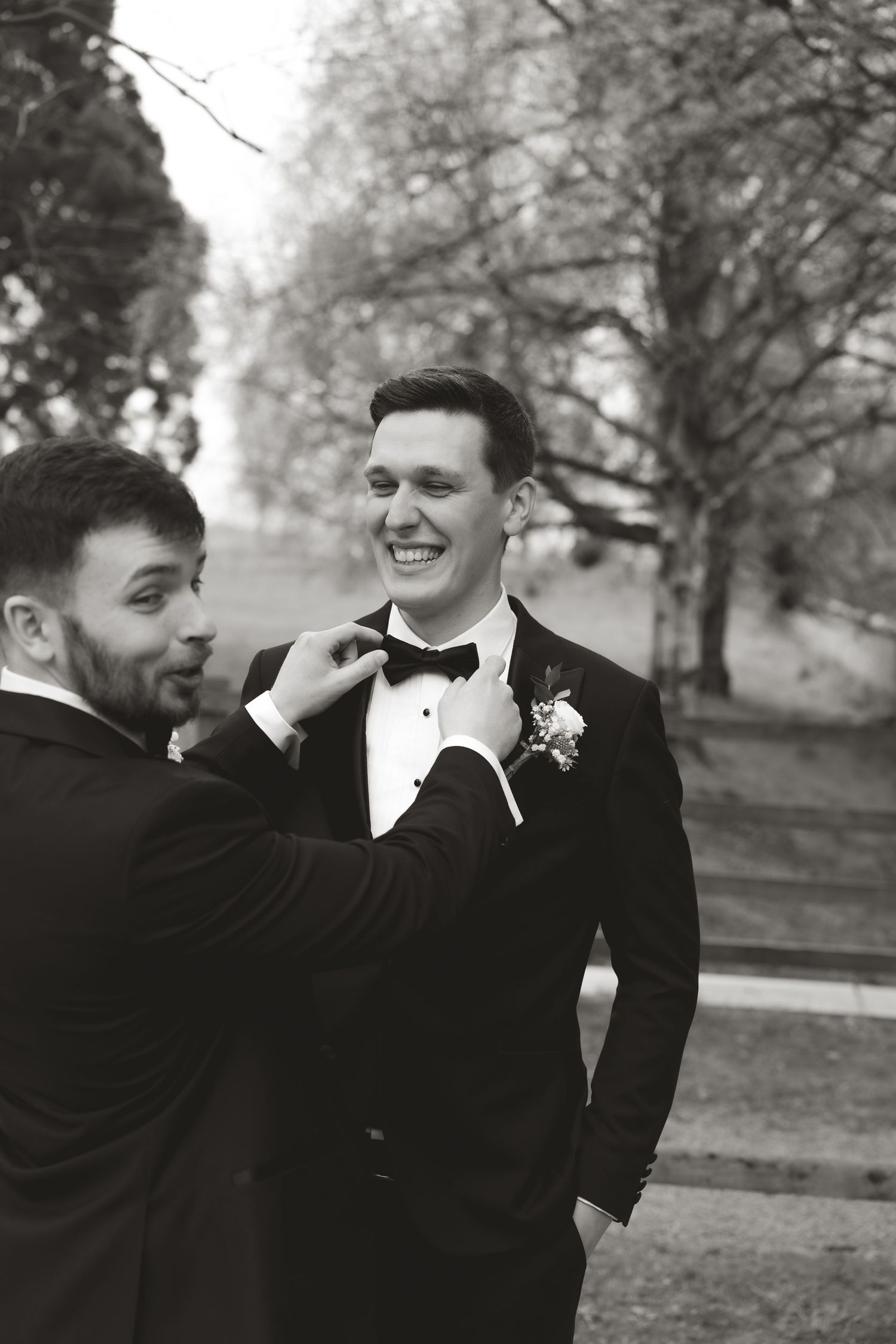 Black and white candid shot of groomsmen adjusting a bow tie before the ceremony.