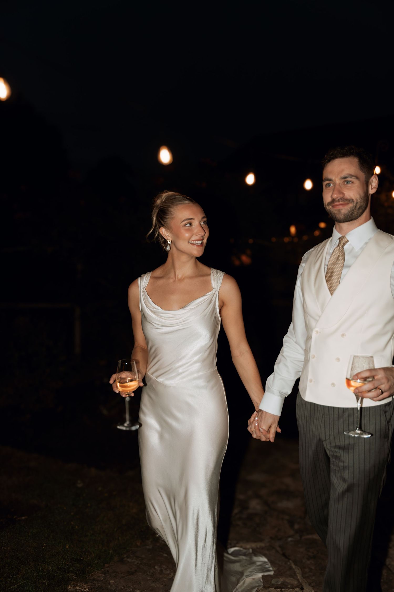 A nighttime celebration with string lights as newlyweds hold champagne glasses.