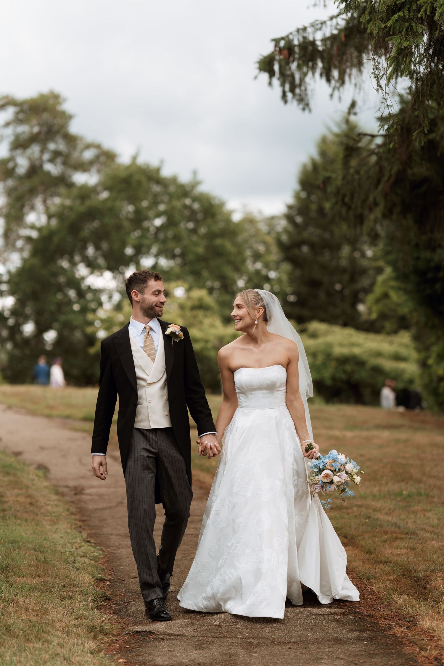 Newlywed couple walking together on a garden path surrounded by trees.
