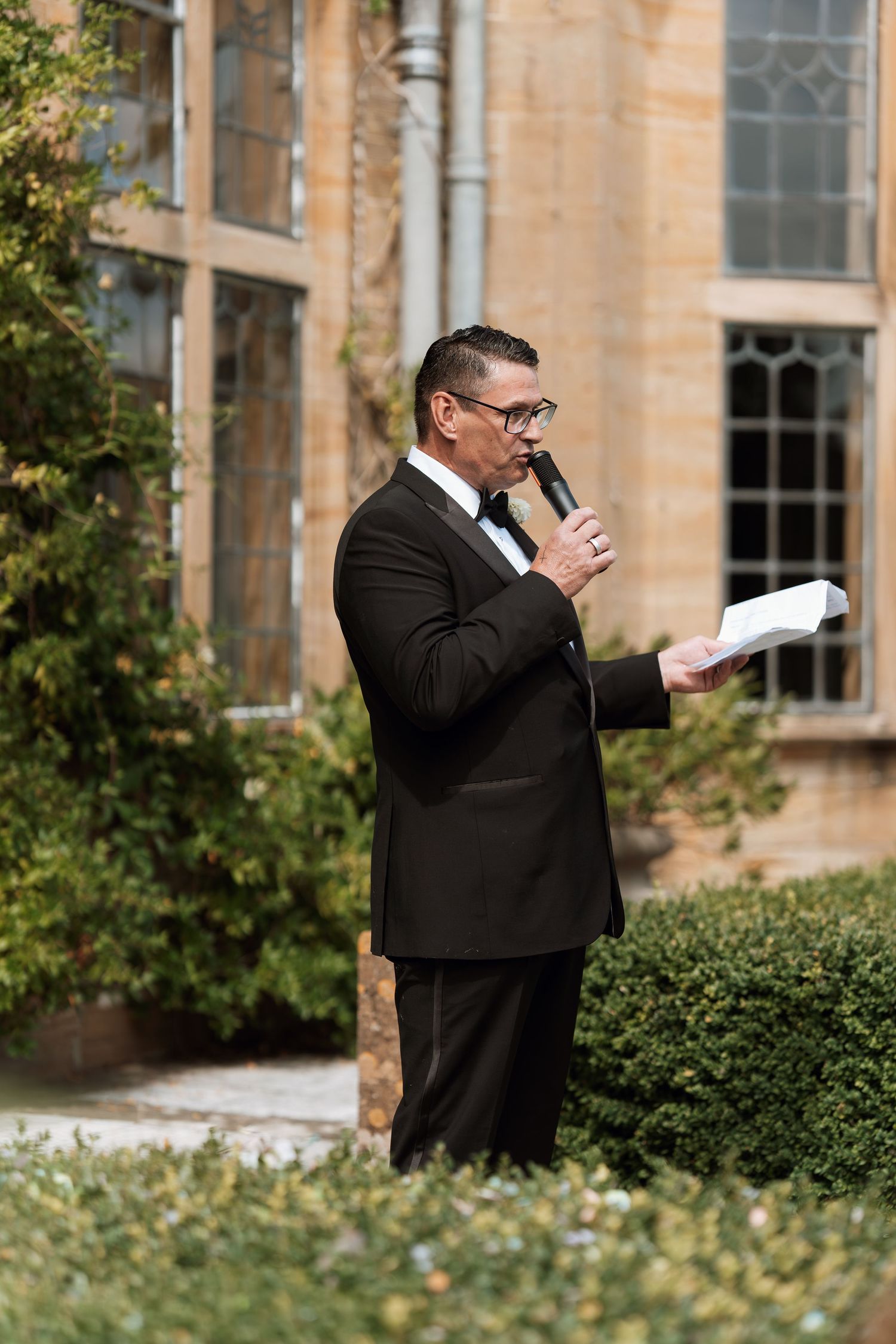 A speaker in a black suit holds a microphone and papers while standing outdoors near a stone building with bushes and windows.