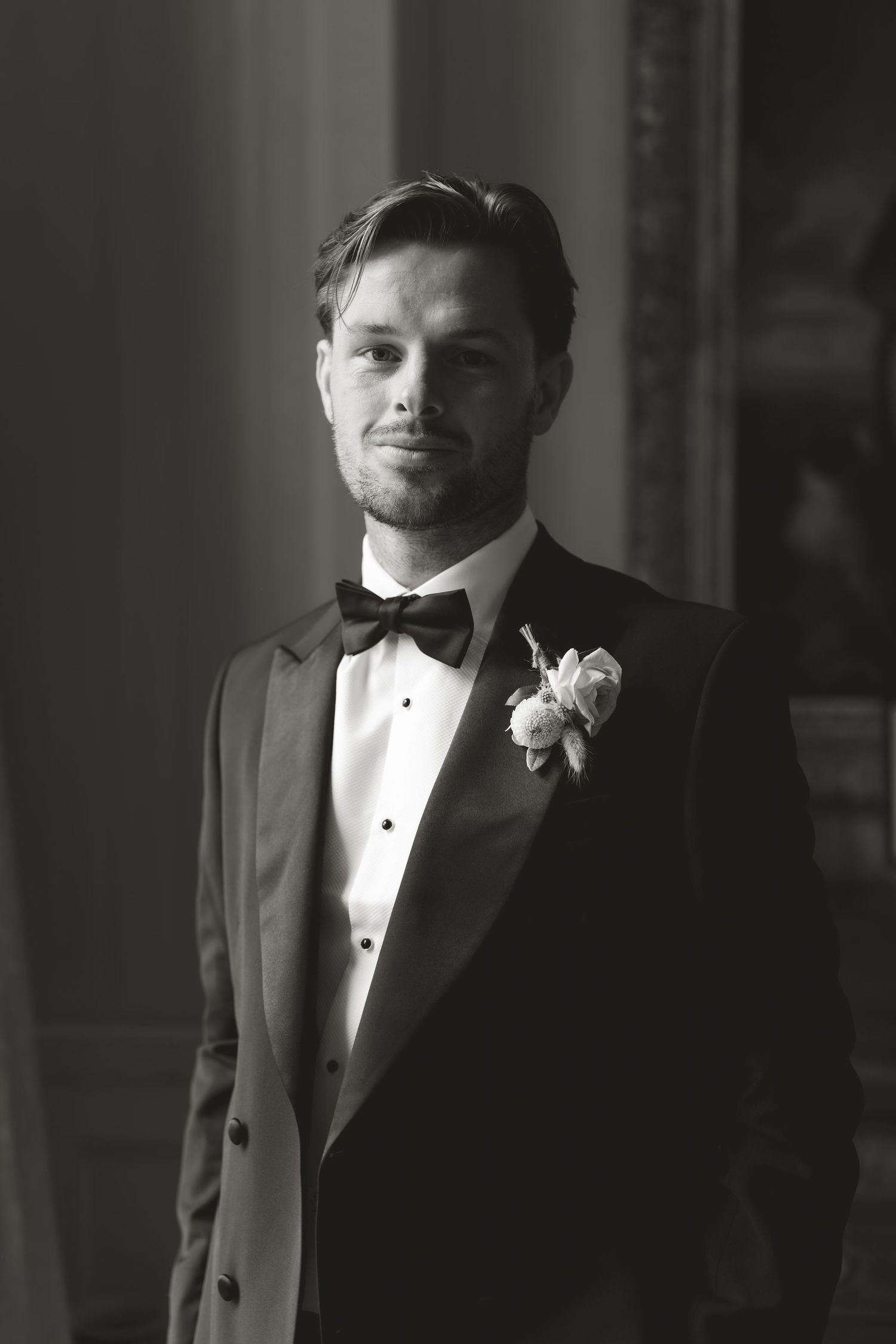 Black and white portrait of a groom in tuxedo with white boutonniere standing in formal pose.