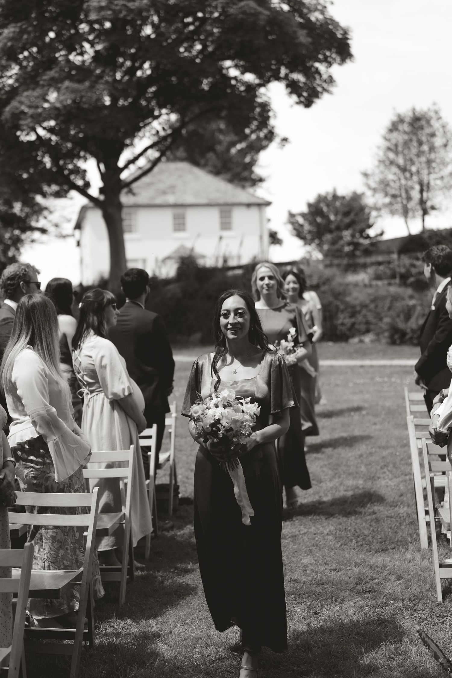 A bridesmaid walks down the aisle at an outdoor wedding ceremony carrying a bouquet.
