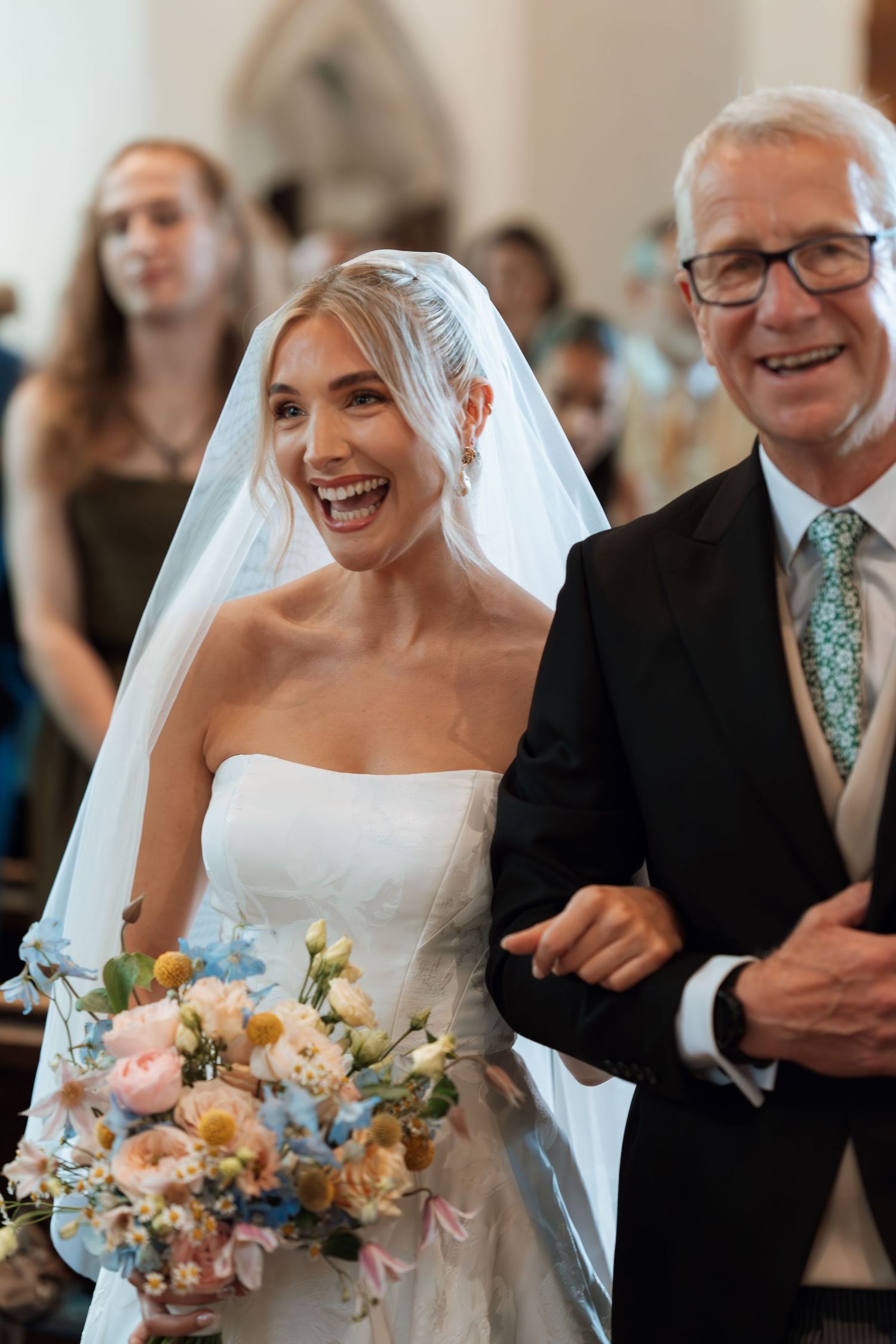 A joyful bride in white dress holds colorful bouquet while walking down church aisle.