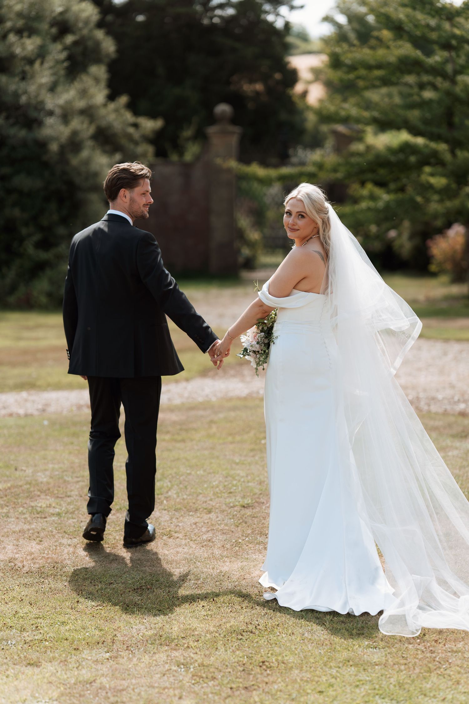 A newlywed couple walking hand in hand through a garden setting with a flowing white wedding dress and veil.