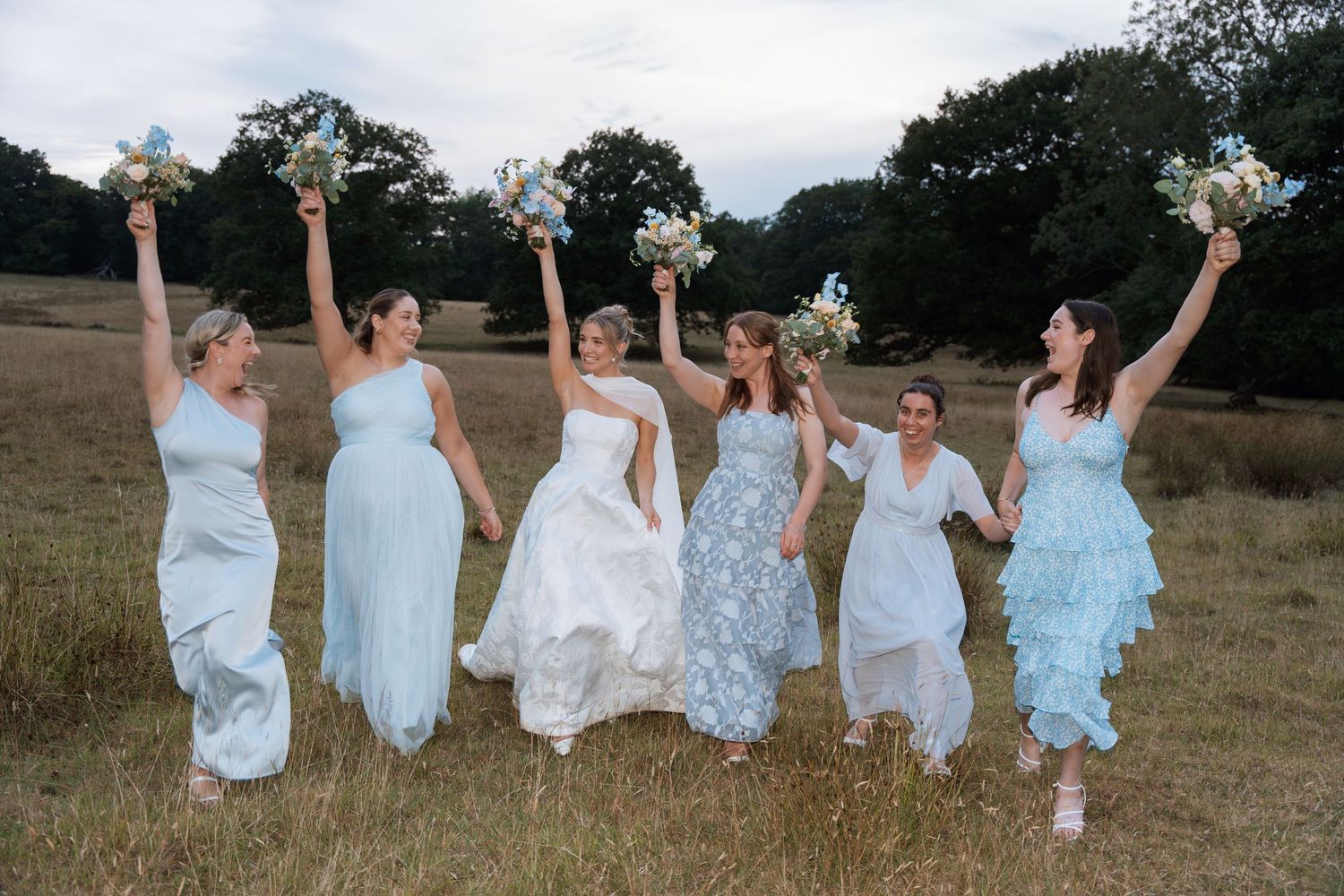 A group wearing light blue and white dresses runs playfully through a grassy field at sunset.