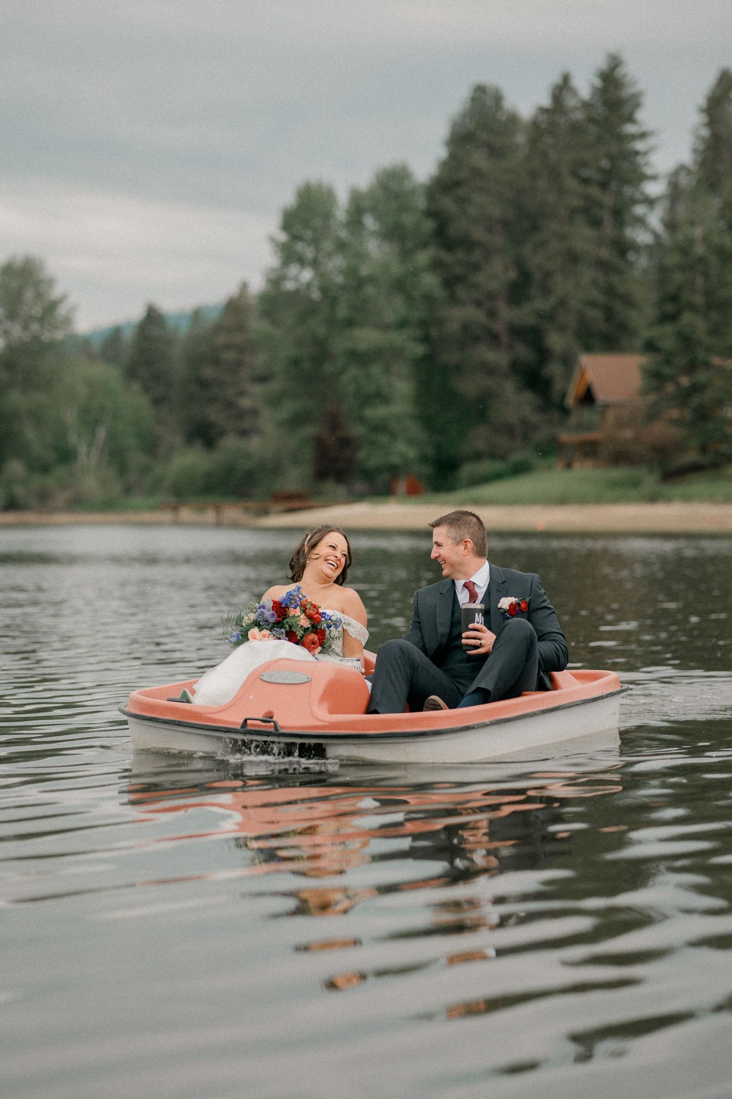 A couple in formal attire share a romantic moment in a pedal boat on a serene lake surrounded by evergreen trees.