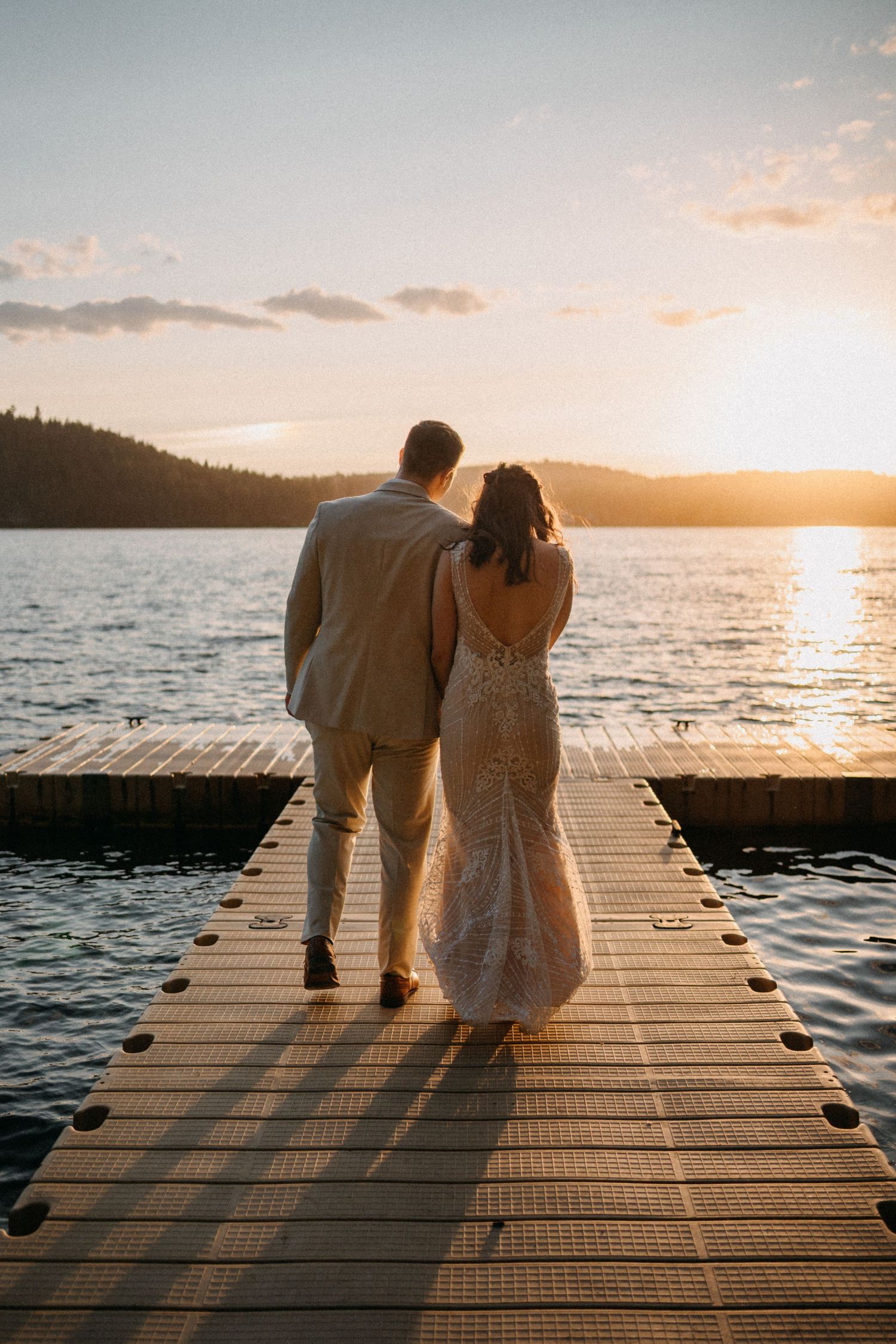 A couple in wedding attire standing on a wooden dock overlooking a lake at sunset.