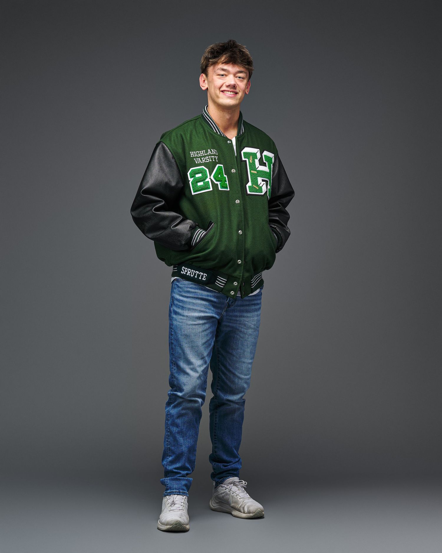 High school senior portrait in green varsity jacket against grey background.