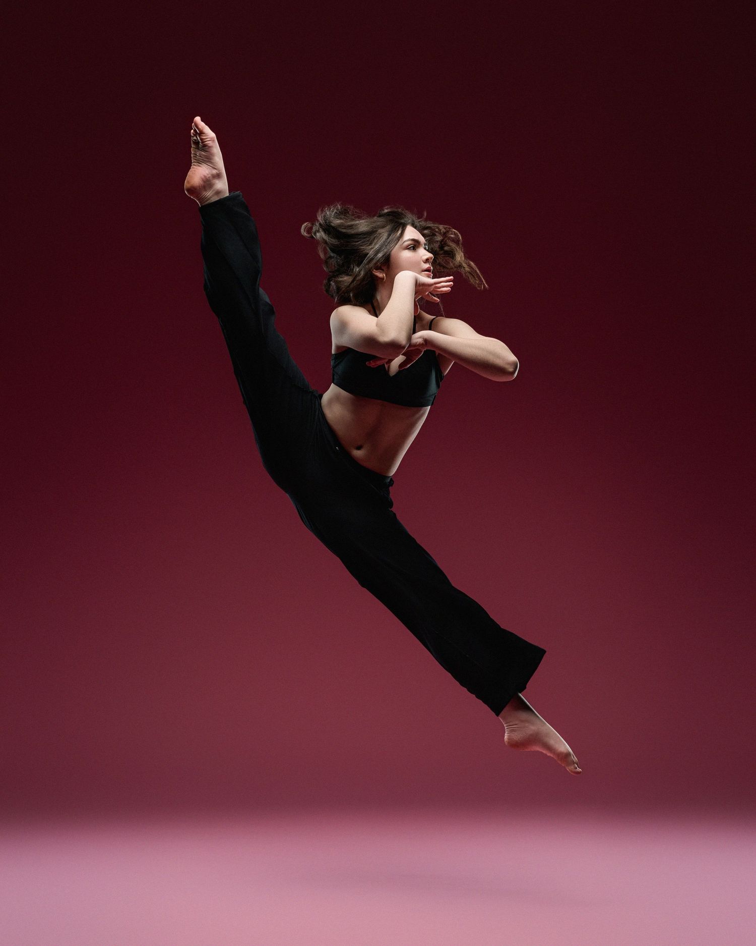 Contemporary dancer performs aerial split jump in black attire against burgundy backdrop.