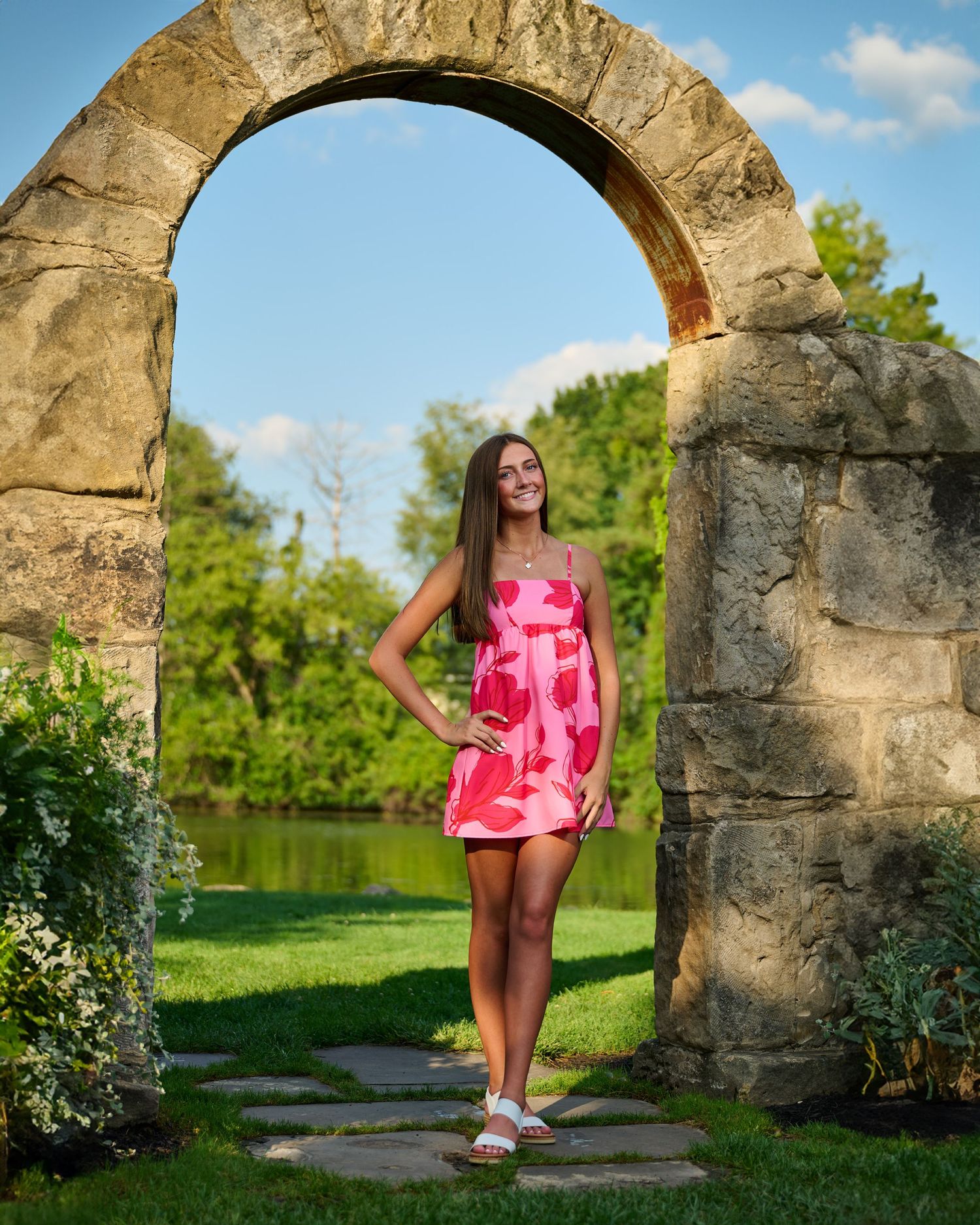 Portrait under stone arch with pink dress and scenic garden background.