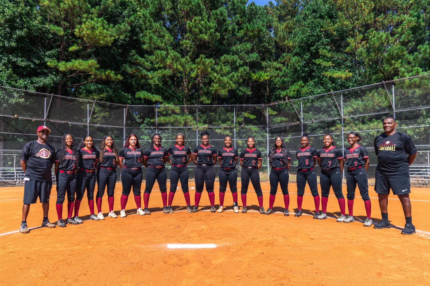 Softball team in navy uniforms poses for group photo on dirt field with trees in background.