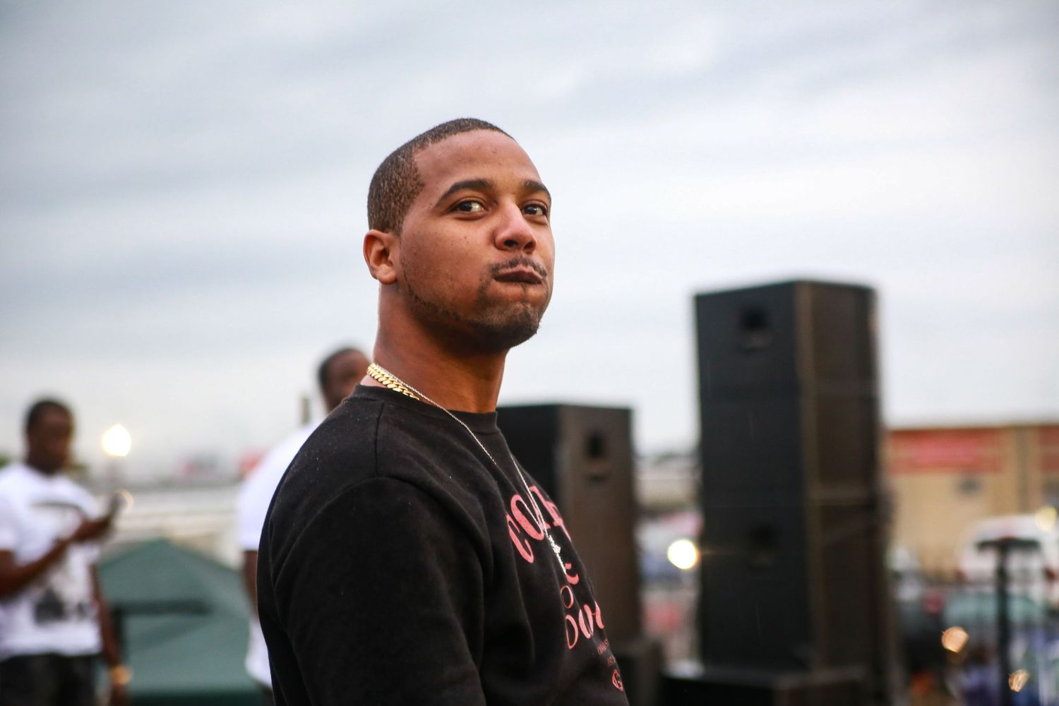 Juelz Santana stands thoughtfully in front of speaker equipment during outdoor concert.