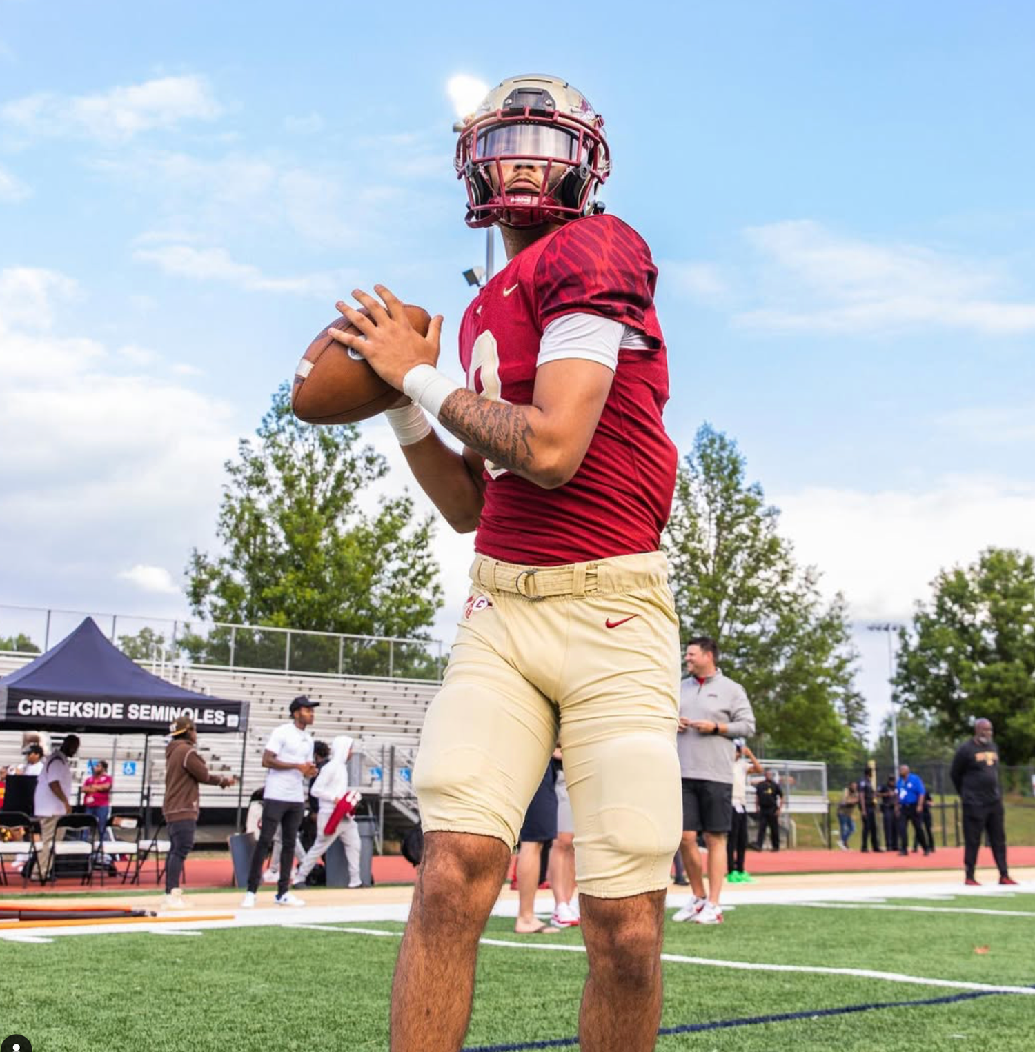 Football player in maroon uniform holds ball in ready position on sunny field.