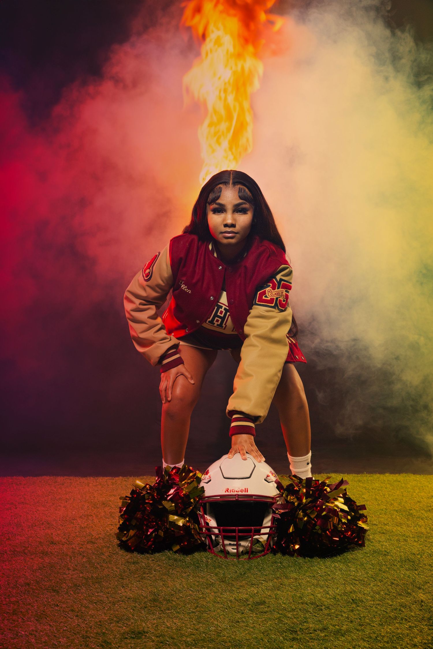 Football varsity jacket wearer poses with helmet and pom poms in colorful smoke.