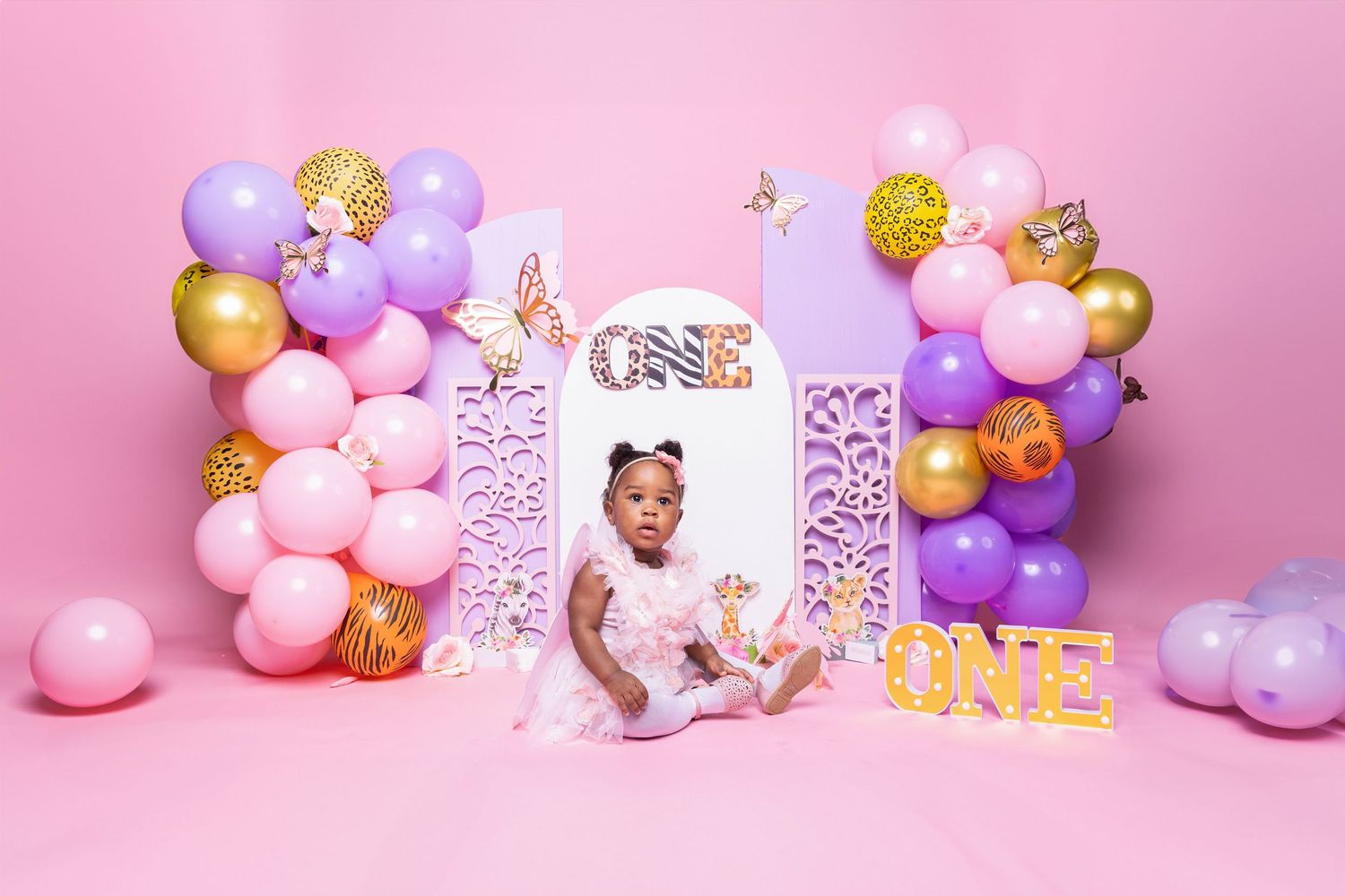 First birthday party setup with pink and purple balloon arch and decorative frame.