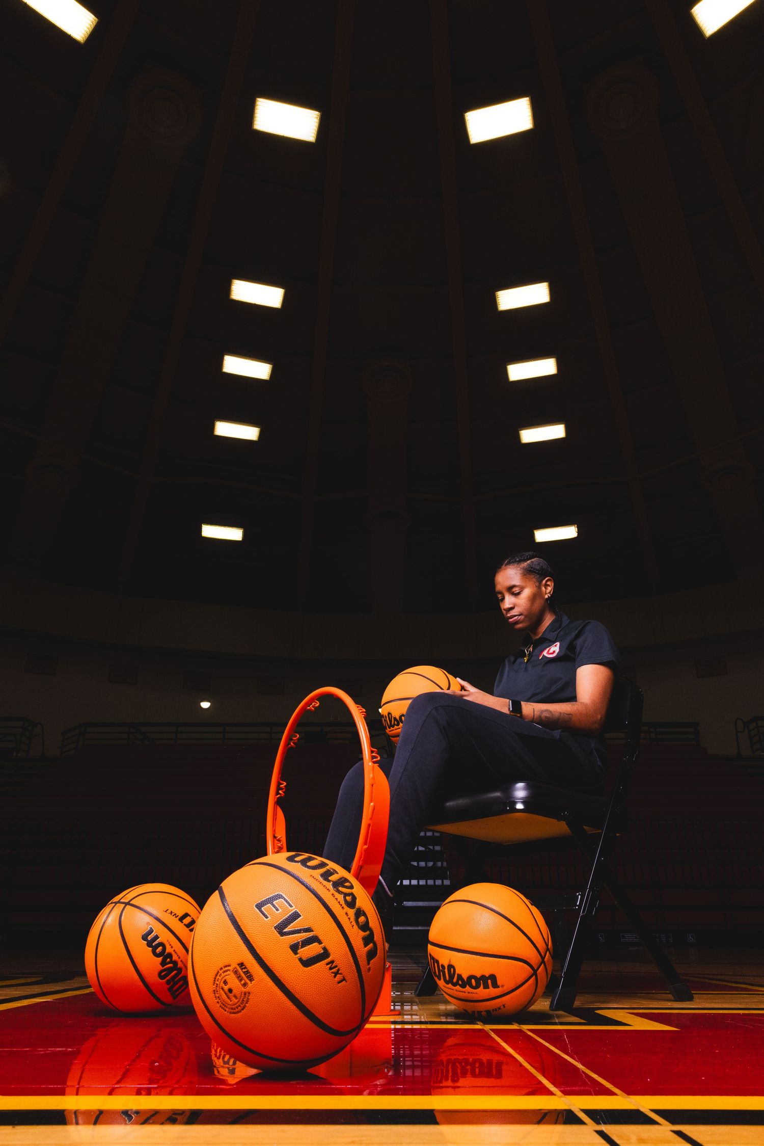 Basketball portrait with player seated among multiple Wilson basketballs in dimly lit gym.
