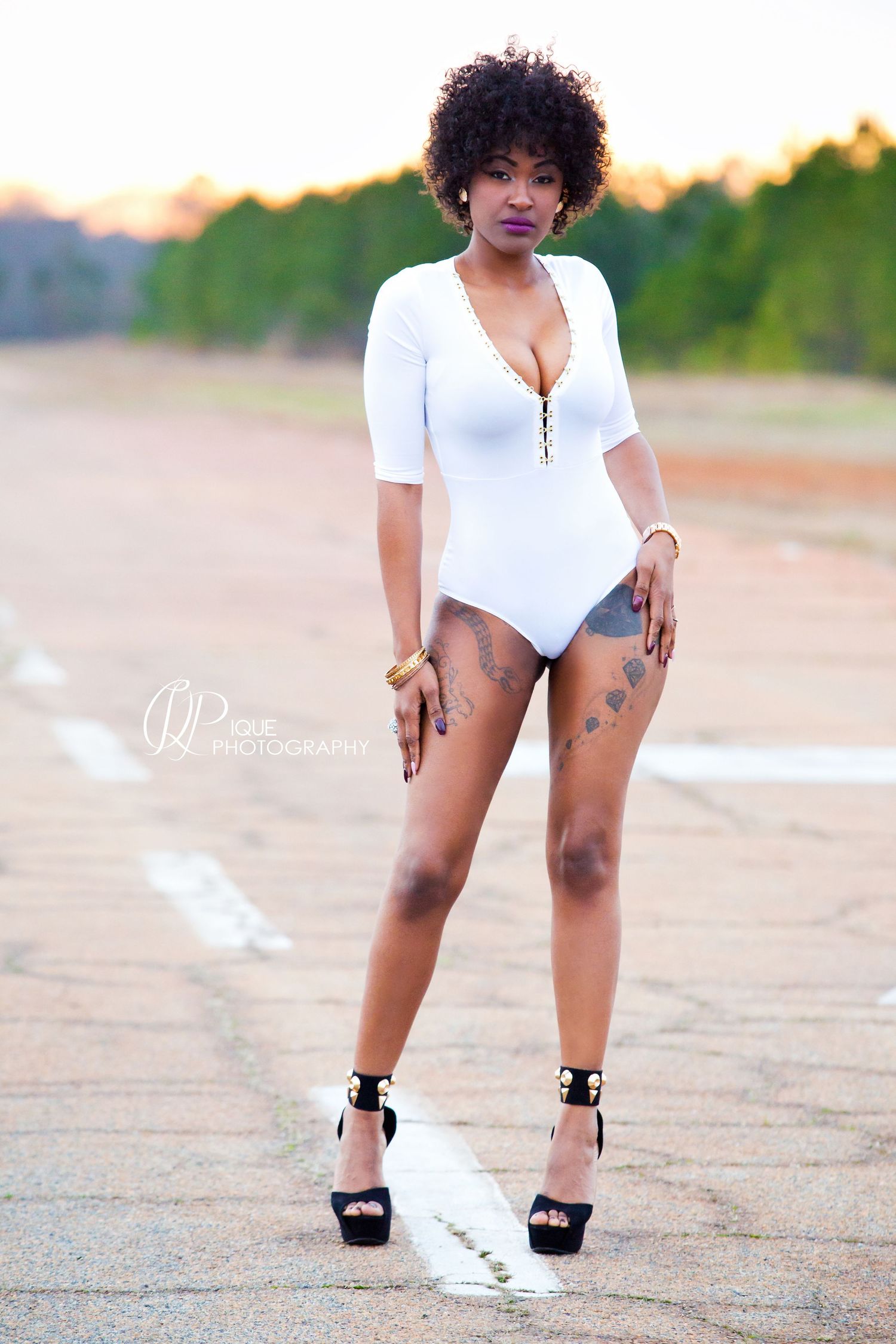 Model in white bodysuit and platform heels poses on outdoor concrete surface.