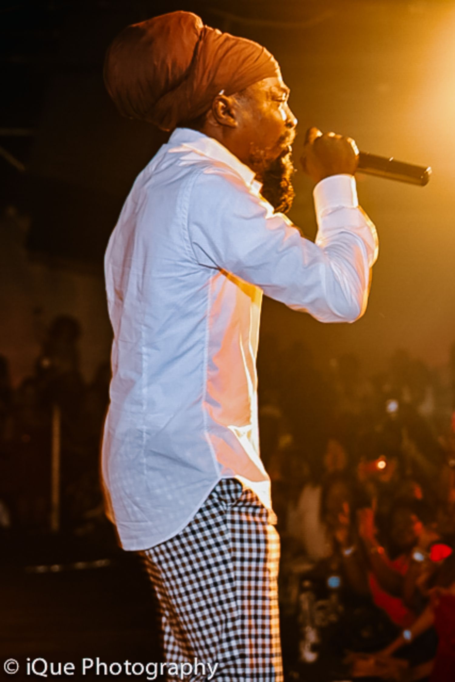Performer in white shirt and checkered pants singing on stage with warm lighting.