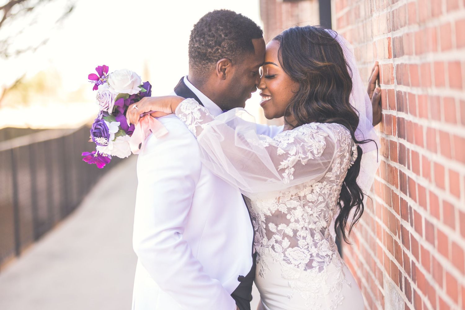 A tender moment between newlyweds against a brick wall with purple floral bouquet.