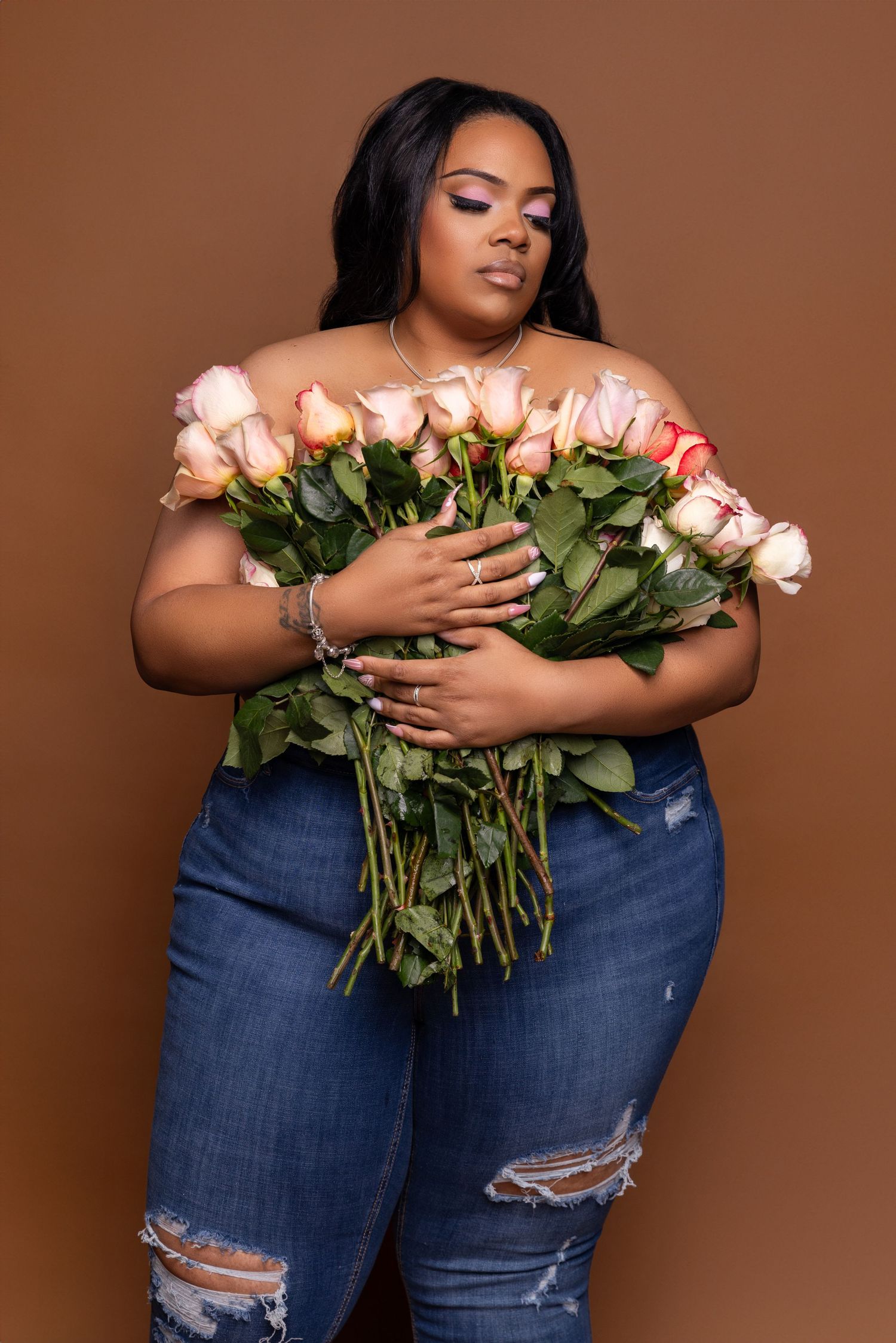 Woman in distressed jeans holding large bouquet of pink roses against brown backdrop.