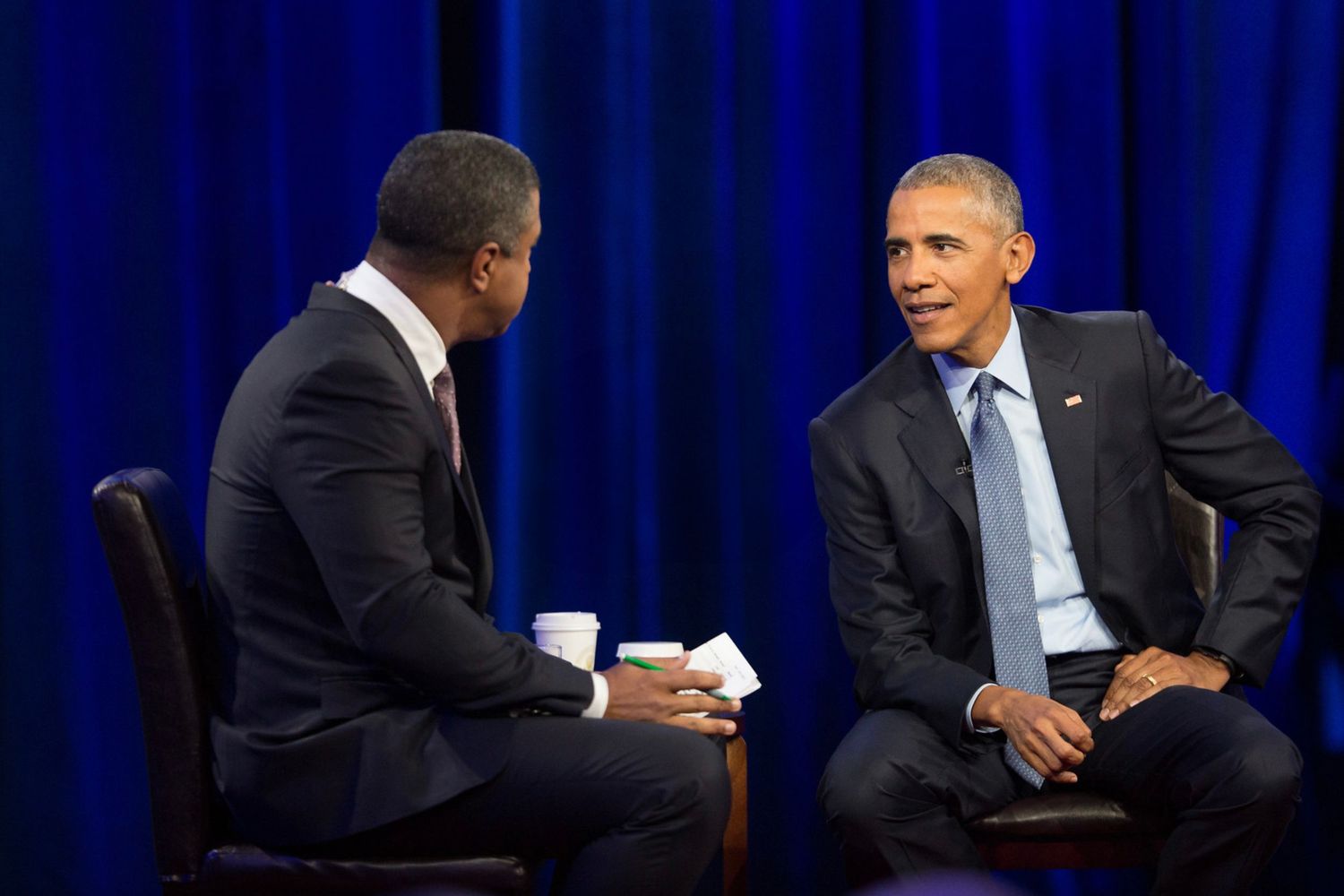 Two figures in suits engage in a televised interview discussion with blue curtains in the background.