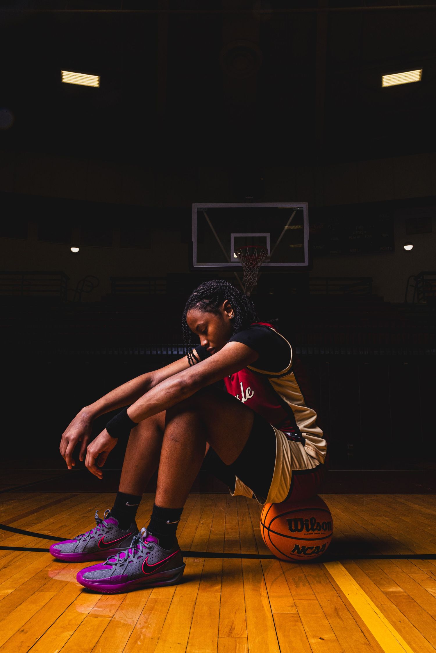 Basketball player in yellow jersey sits on court next to ball in moody gym lighting.