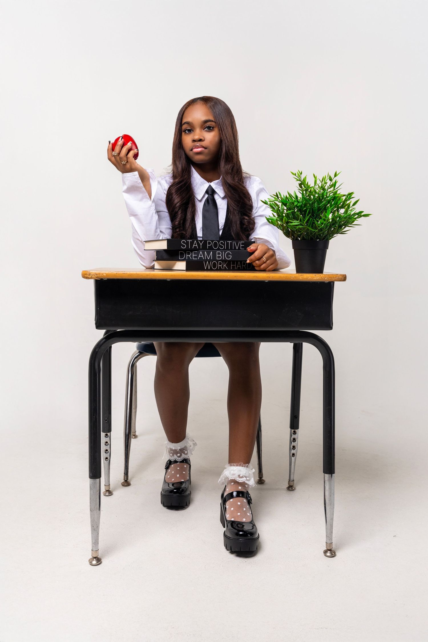 Student at desk holding red apple with nameplate, plant and wearing school uniform in studio setting.