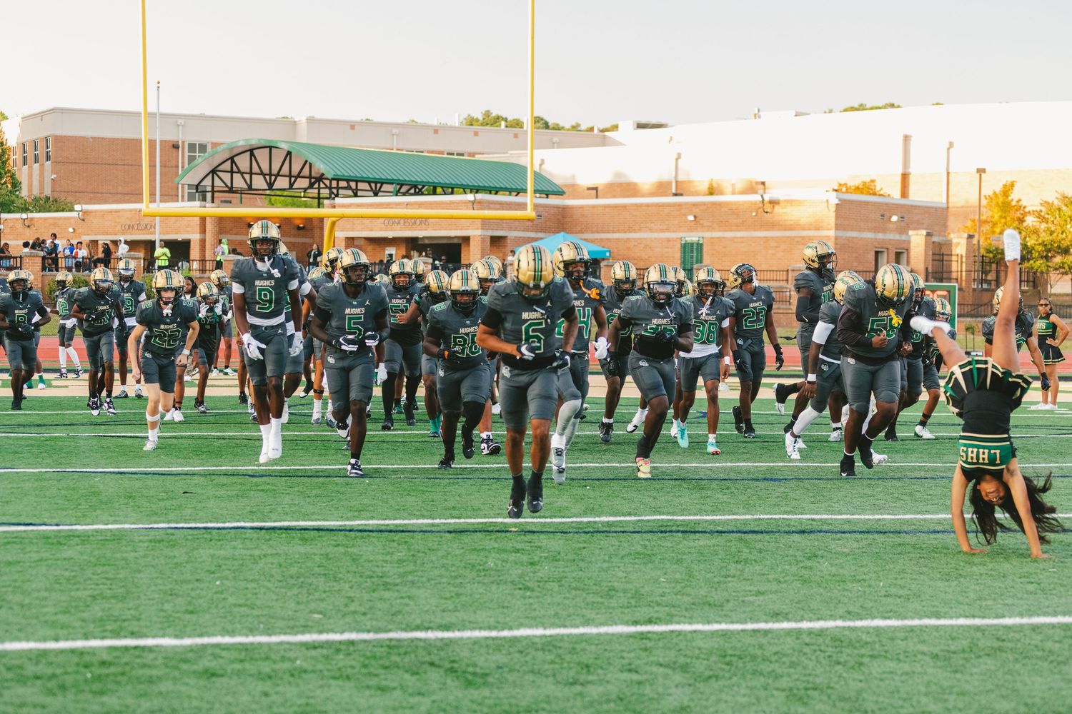 High school football players practice drills on a green turf field during sunset with coaches and players visible.