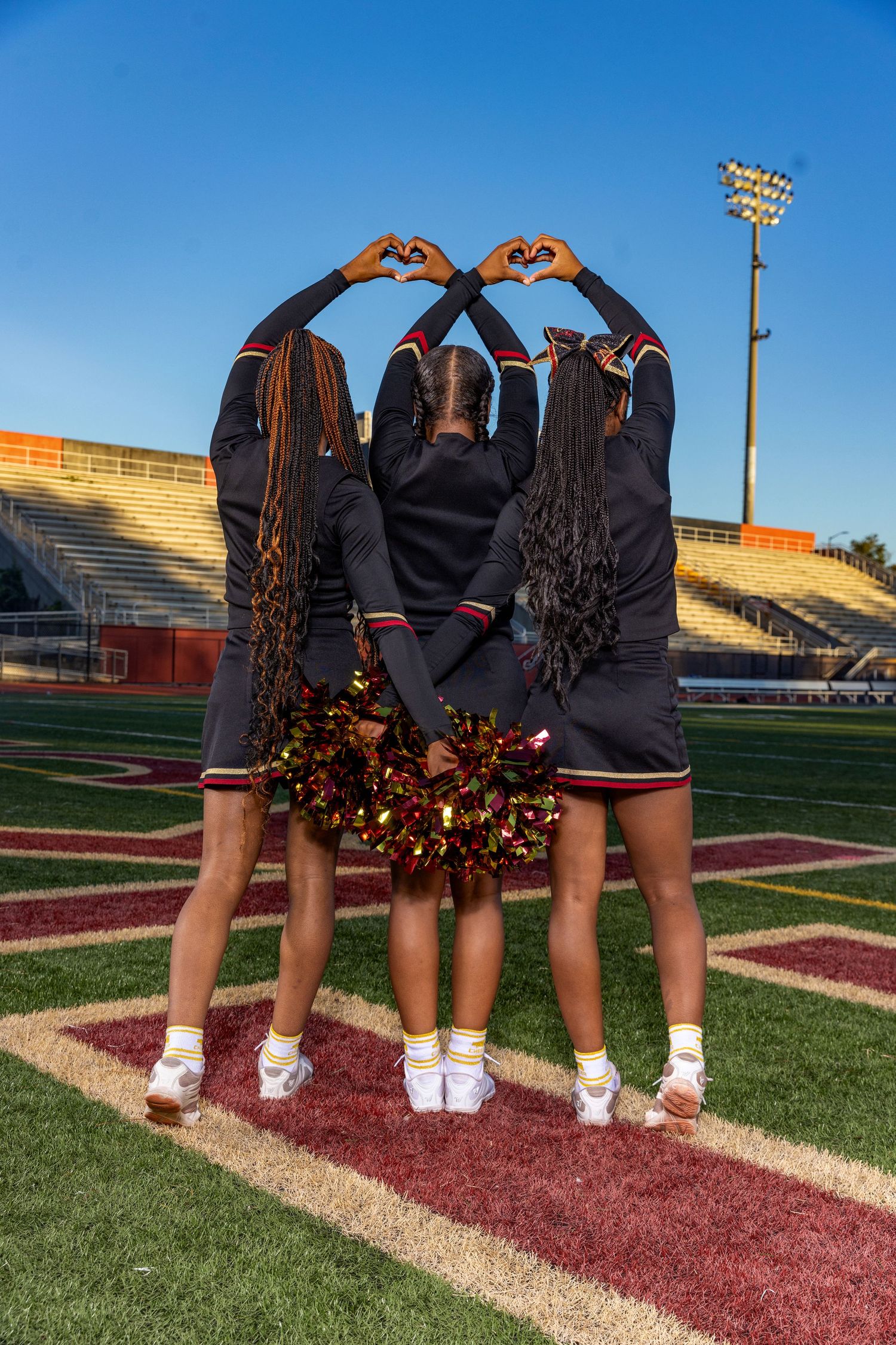Three cheerleaders make heart shapes with hands while facing away on football field.