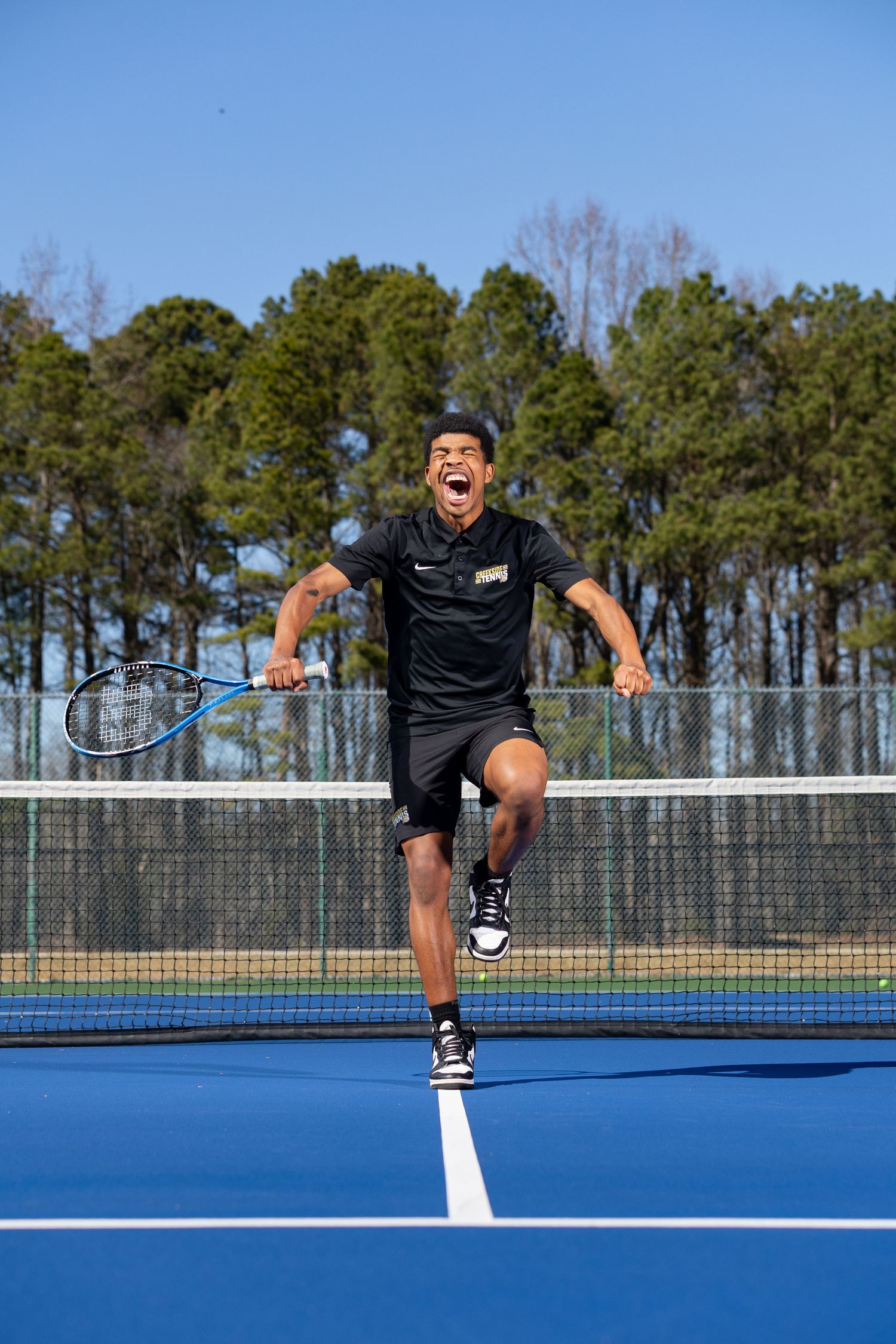 Tennis player leaps with racquet on blue court against pine tree background.