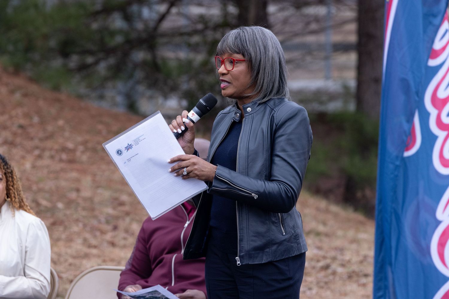 Speaker holding microphone at outdoor event with presentation board visible.