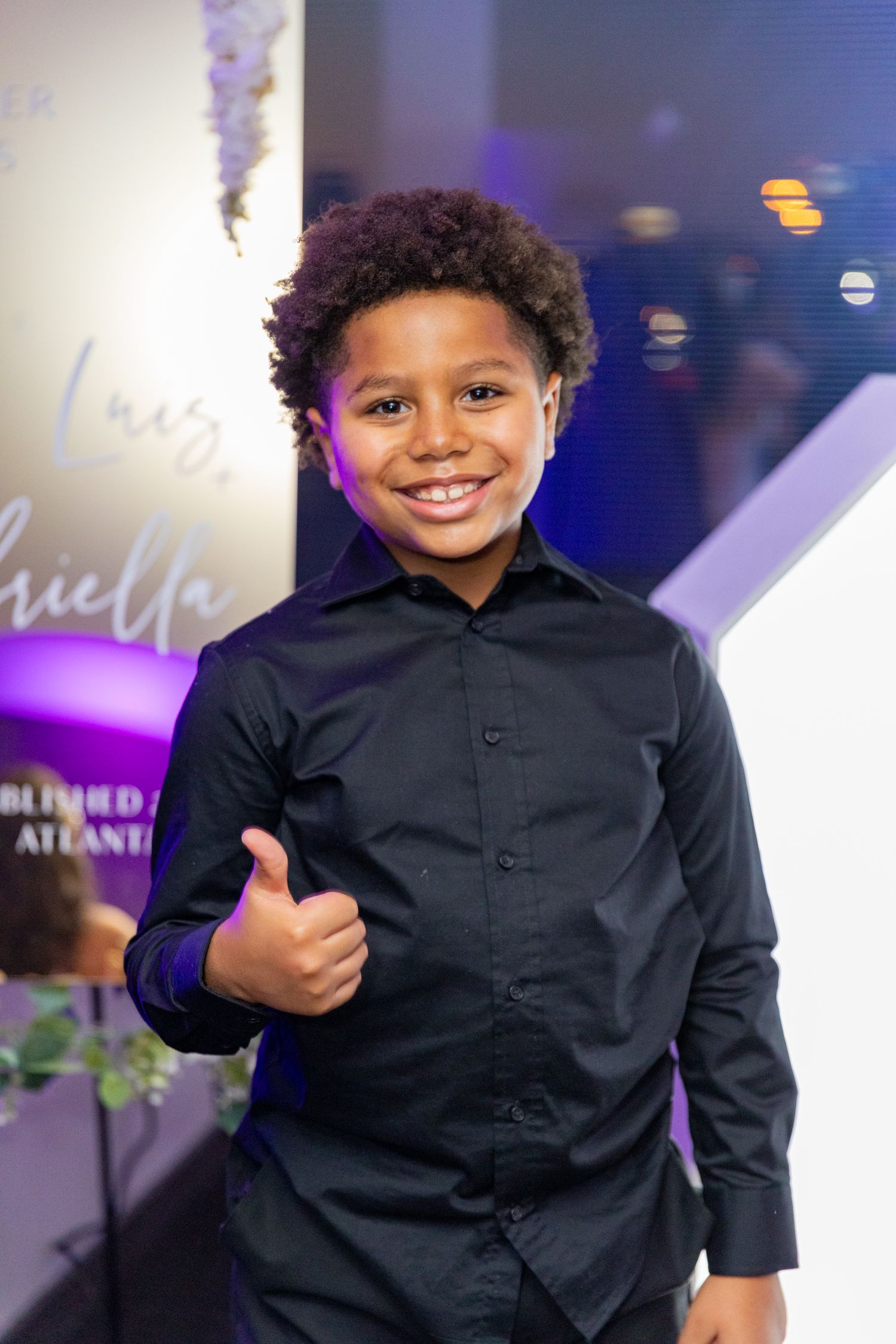 Young child in black formal attire giving thumbs up at stylish event with purple lighting.