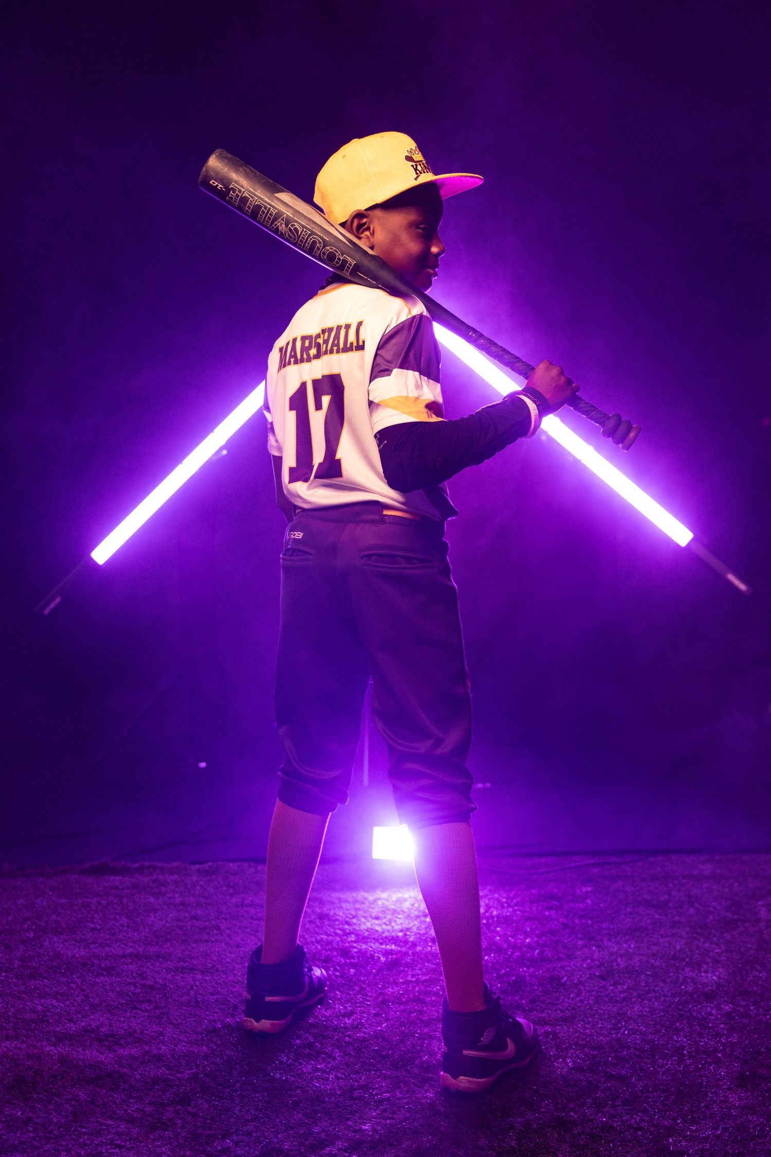 Youth baseball player poses with light-up neon tubes against purple background.