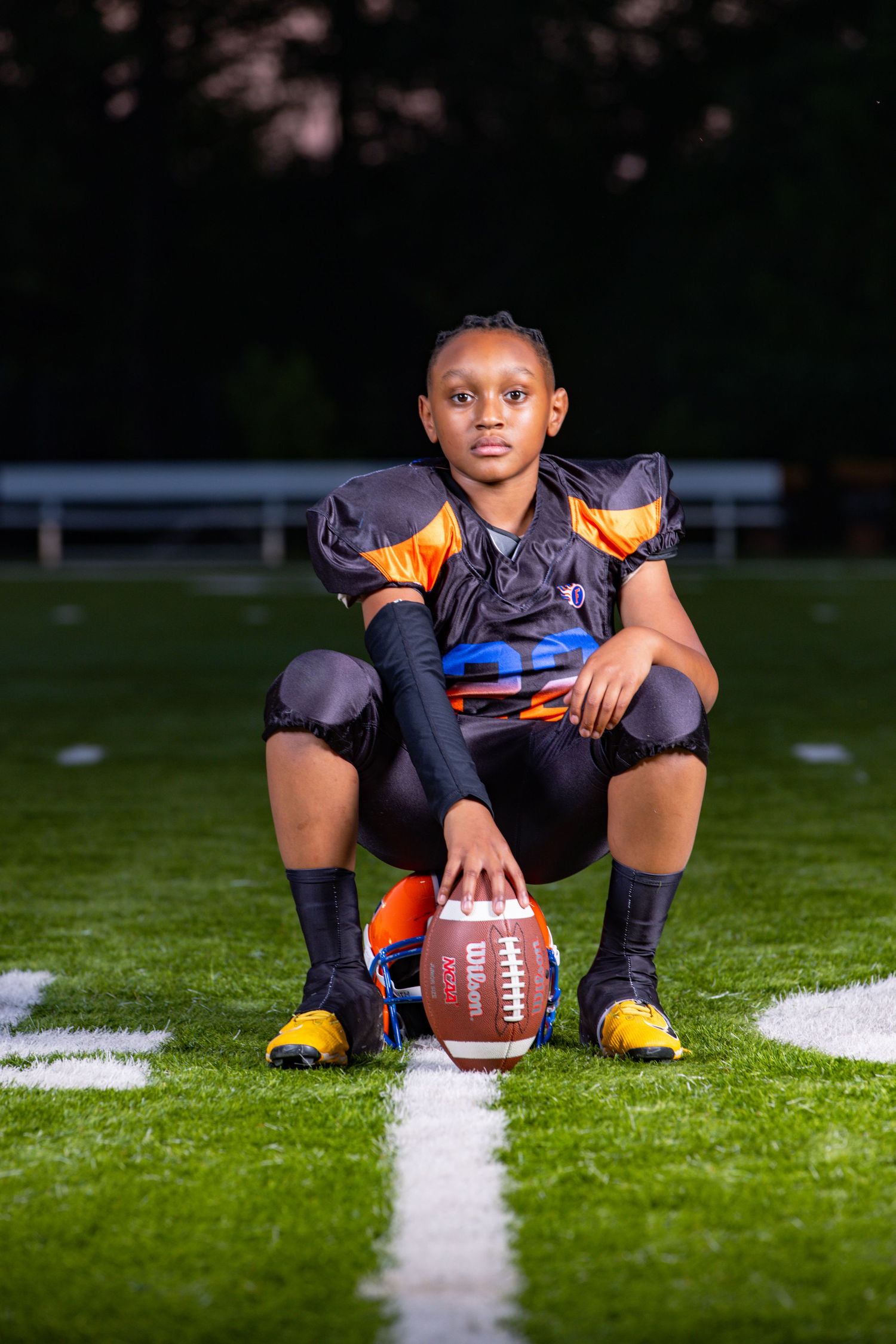 A youth football player in orange and navy uniform sits on a football field with a ball.