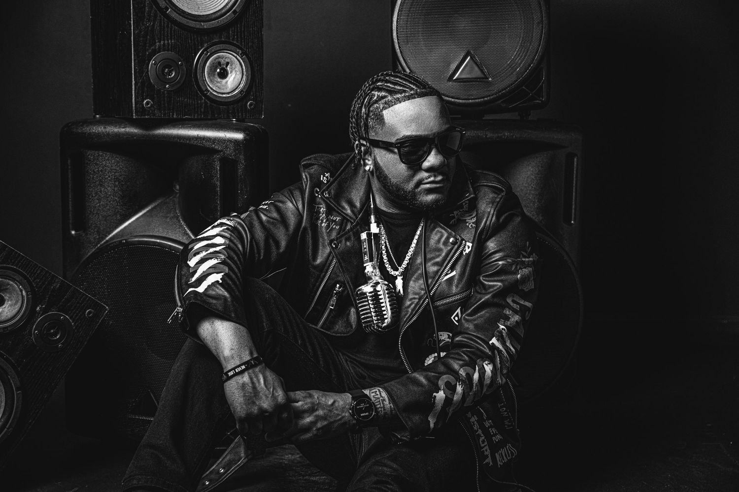 Black and white portrait of musician in leather jacket seated among studio speakers.