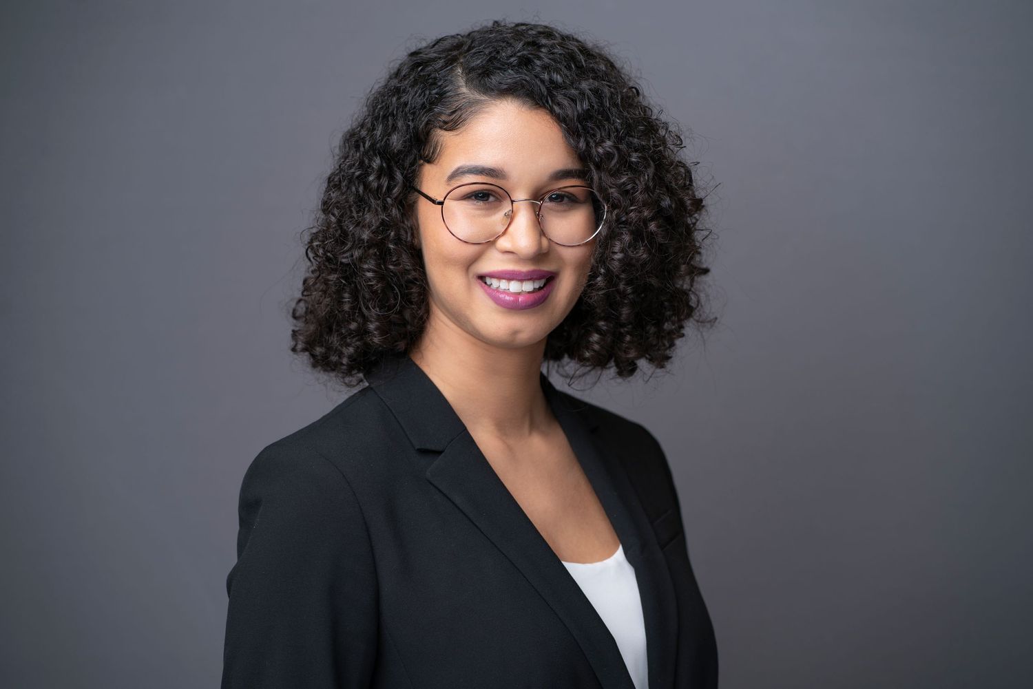 Professional portrait of businesswoman with curly hair and glasses against gray backdrop.