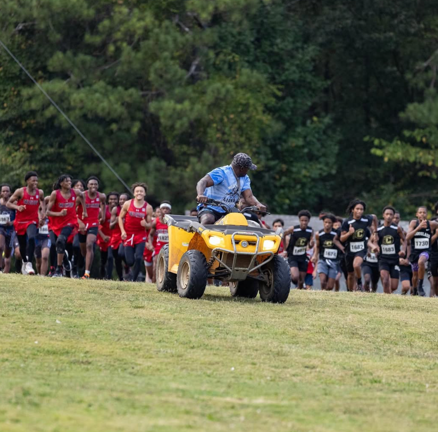 ATV leads crowd of runners during cross country race on grassy hill.