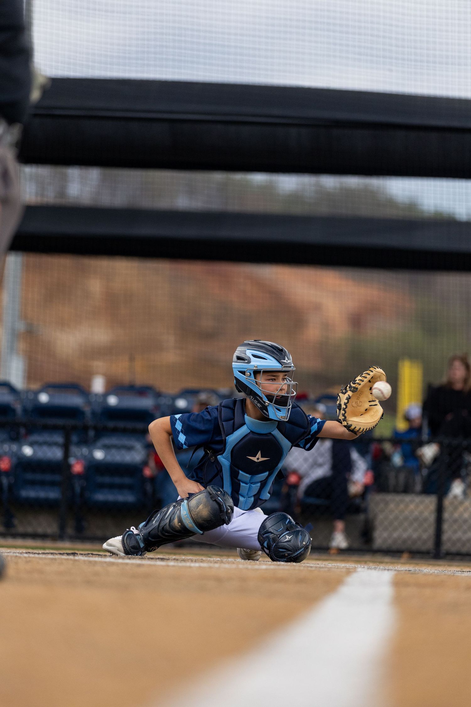 Baseball catcher in light blue uniform crouching behind home plate in ready position.