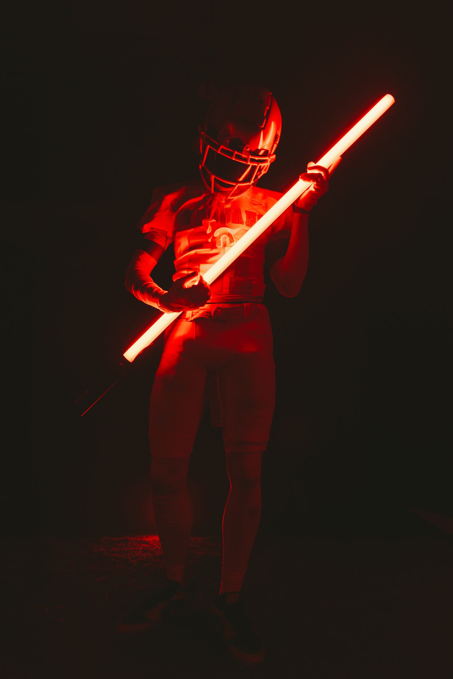 Athlete in red football uniform holds glowing red light staff in dark atmospheric setting.