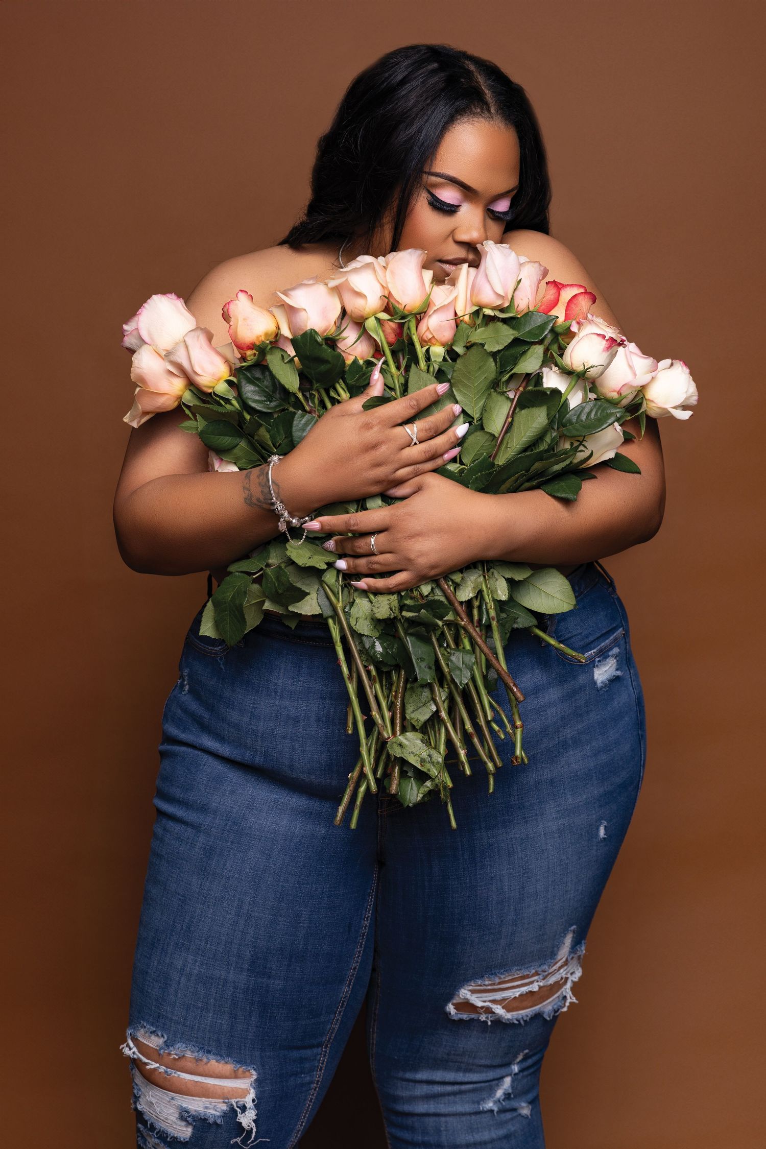 Fashion model embracing pink roses while wearing distressed denim against brown background.