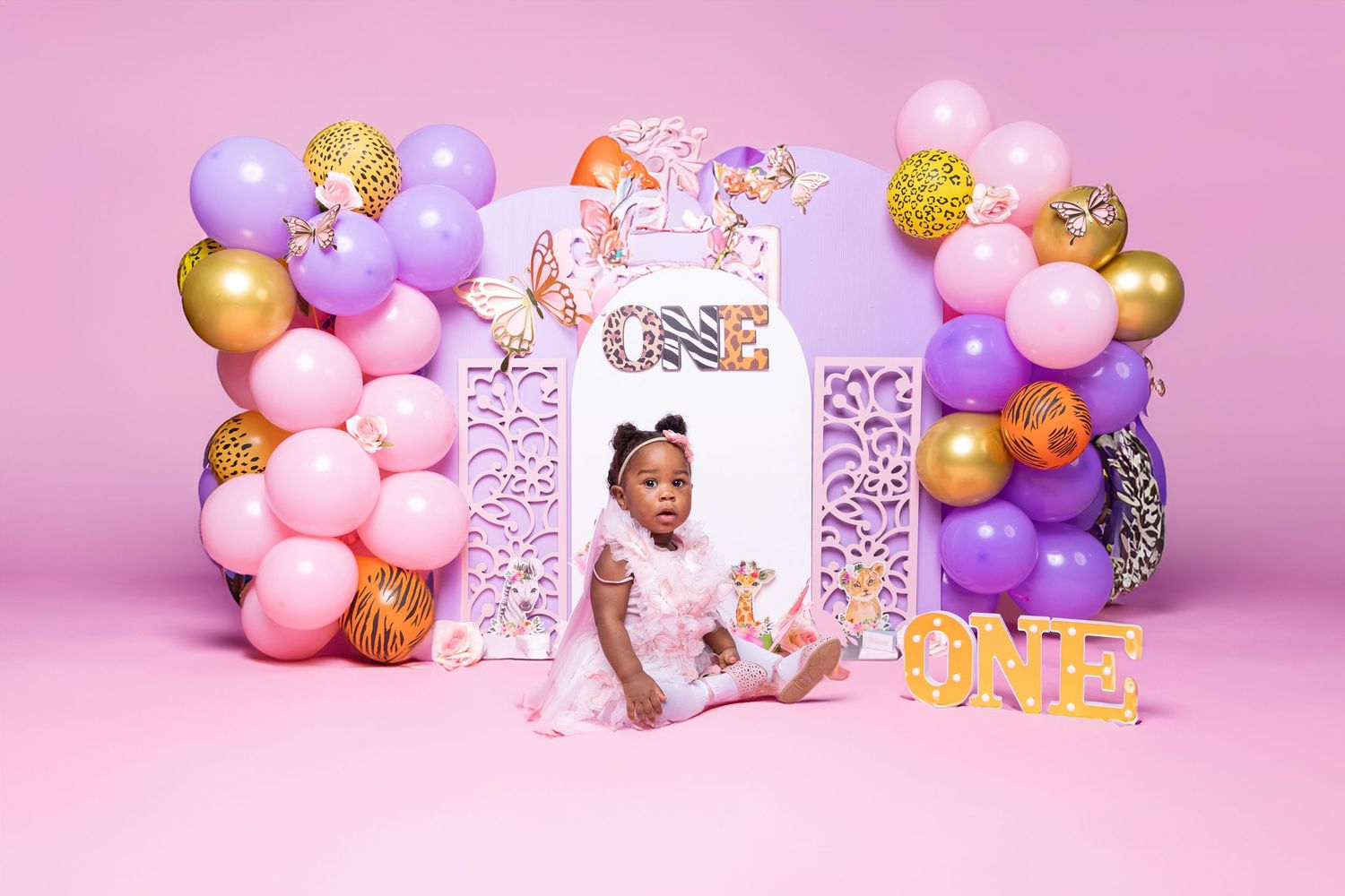 First birthday party setup with pink and purple balloon arch and decorative letters against pink backdrop.