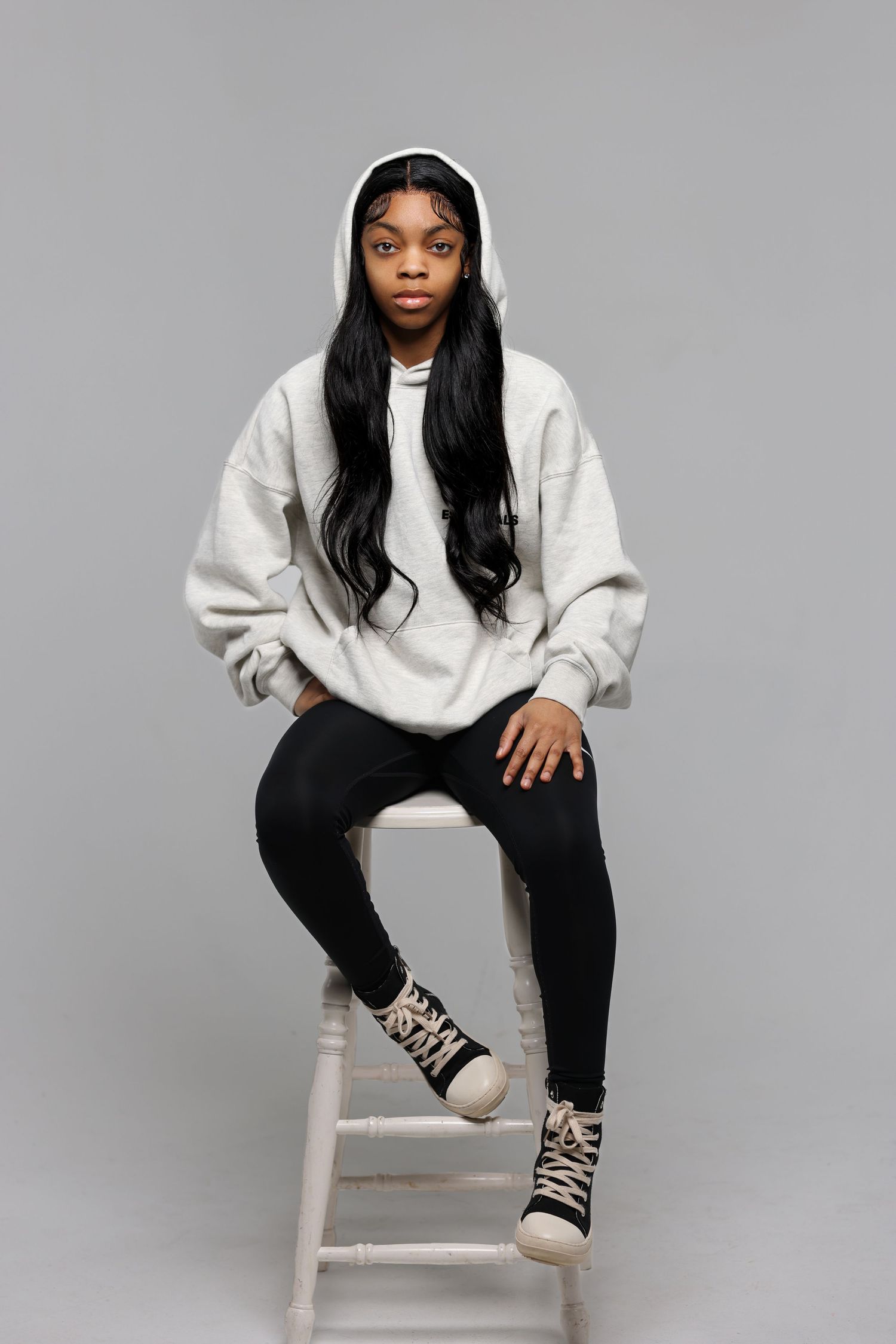 Model in casual white sweater and black pants poses on white stool against gray background.